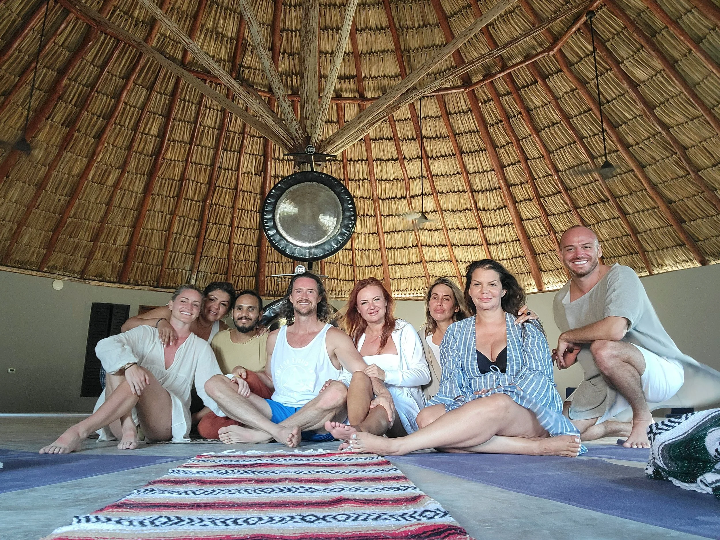 A group of nine people sitting on yoga mats in a yoga studio with a thatched roof and a large gong hanging from the ceiling.