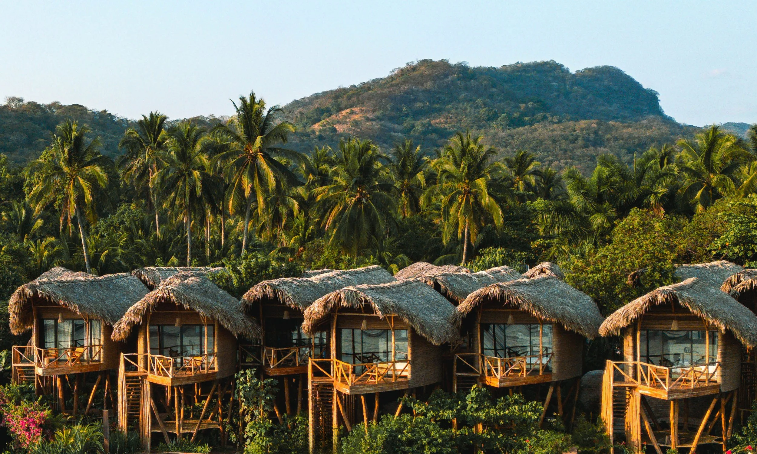 Row of thatched-roof overwater bungalows on stilts surrounded by lush greenery and tall palm trees with mountains in the background.