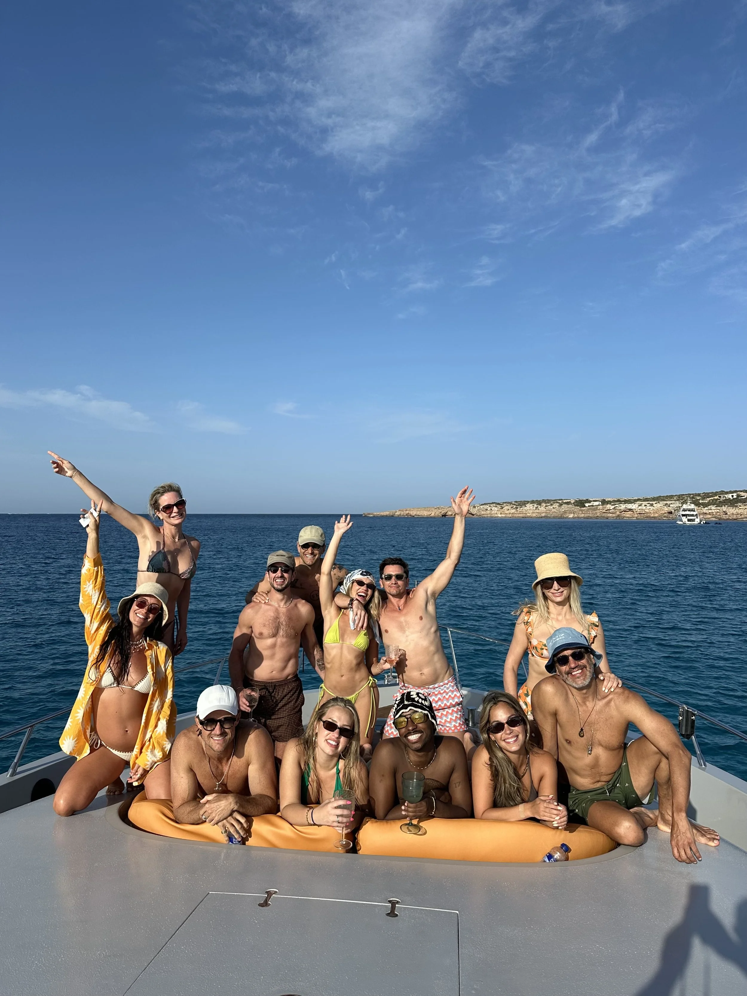A group of people, dressed in swimwear, are enjoying a sunny day on a boat in the ocean whilst on a retreat, with some smiling, waving, and holding drinks, against a backdrop of clear blue skies and water.