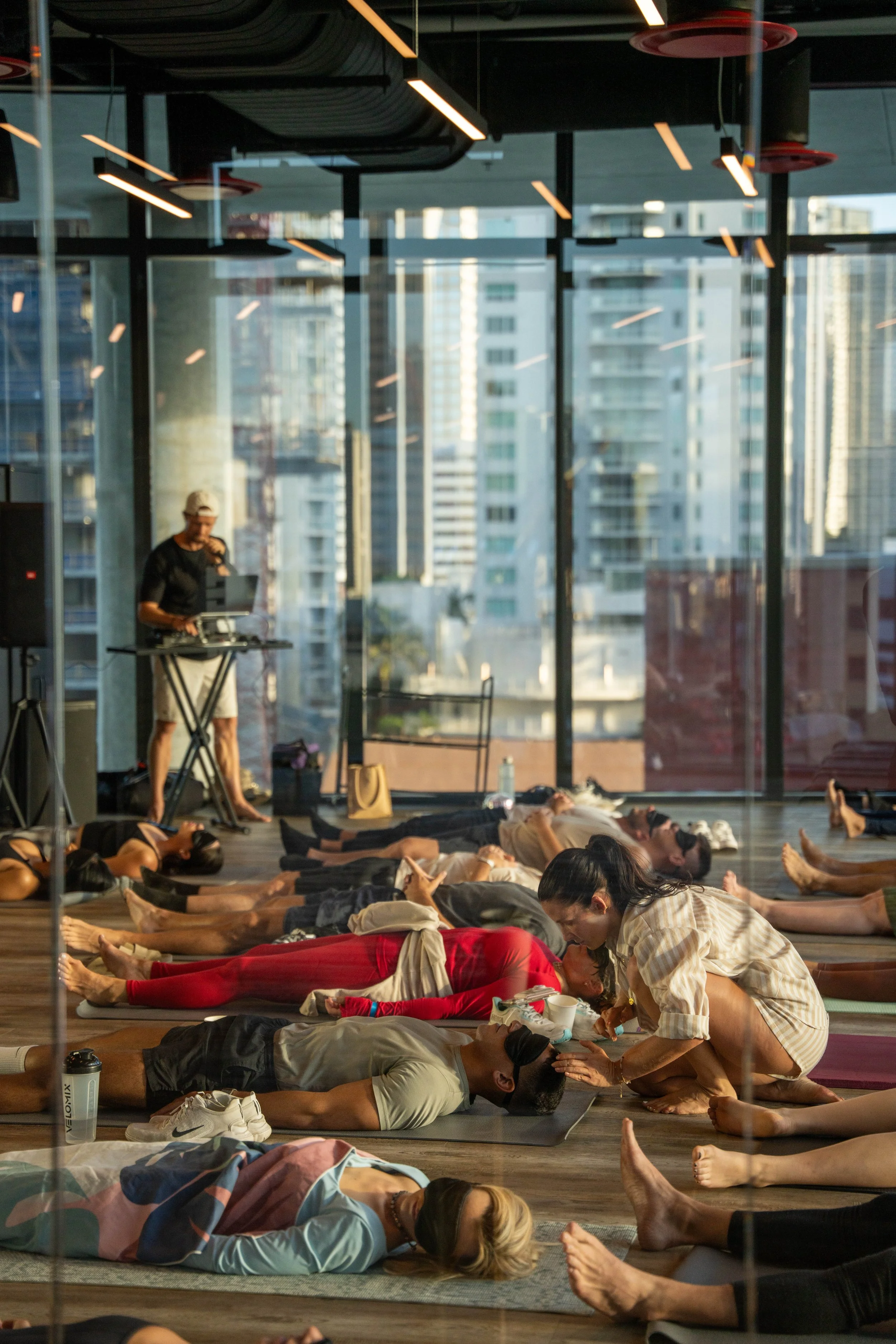 A group of people participating in a yoga or meditation class in a sunlit studio with large windows and a cityscape view, while an instructor offers guidance.