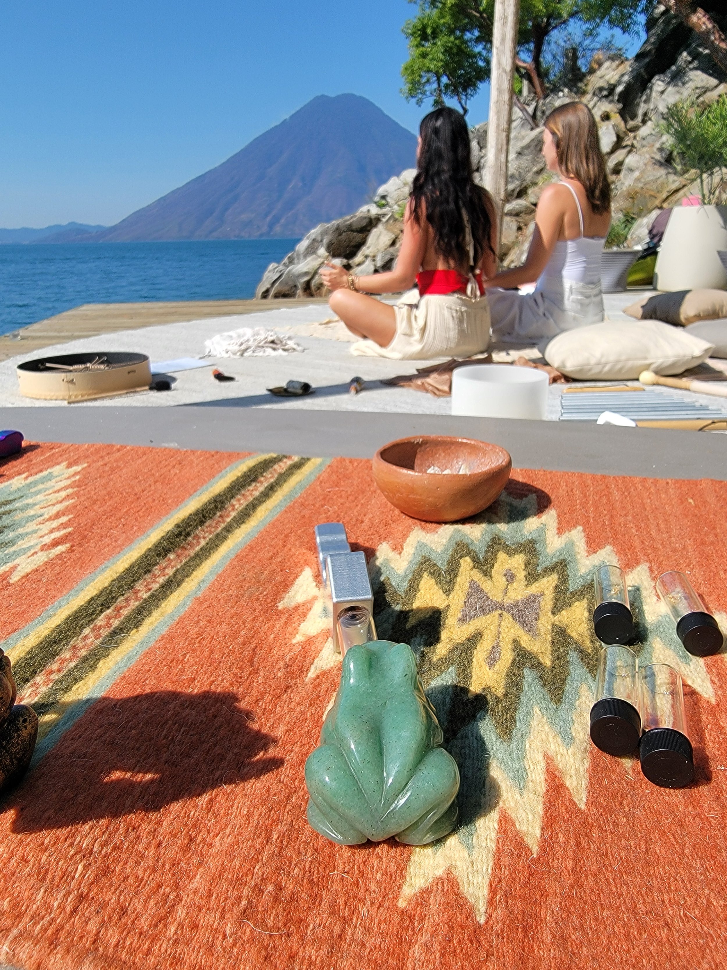 Two women sitting on a mat by a lake with a volcano in the background, surrounded by trees and rocks, during a sunny day. The foreground shows a colorful rug with a frog-shaped object, a small bowl, and makeup or perfume bottles.
