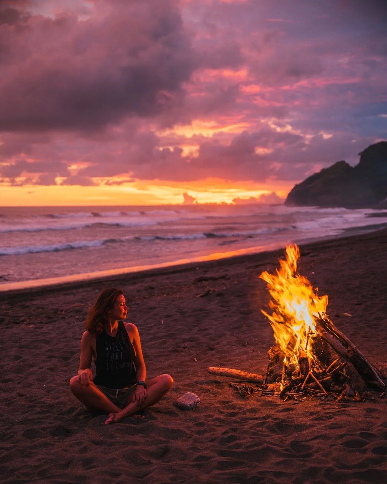 A woman sitting cross-legged on a sandy beach at sunset near a campfire with ocean waves and a cloudy sky in the background.
