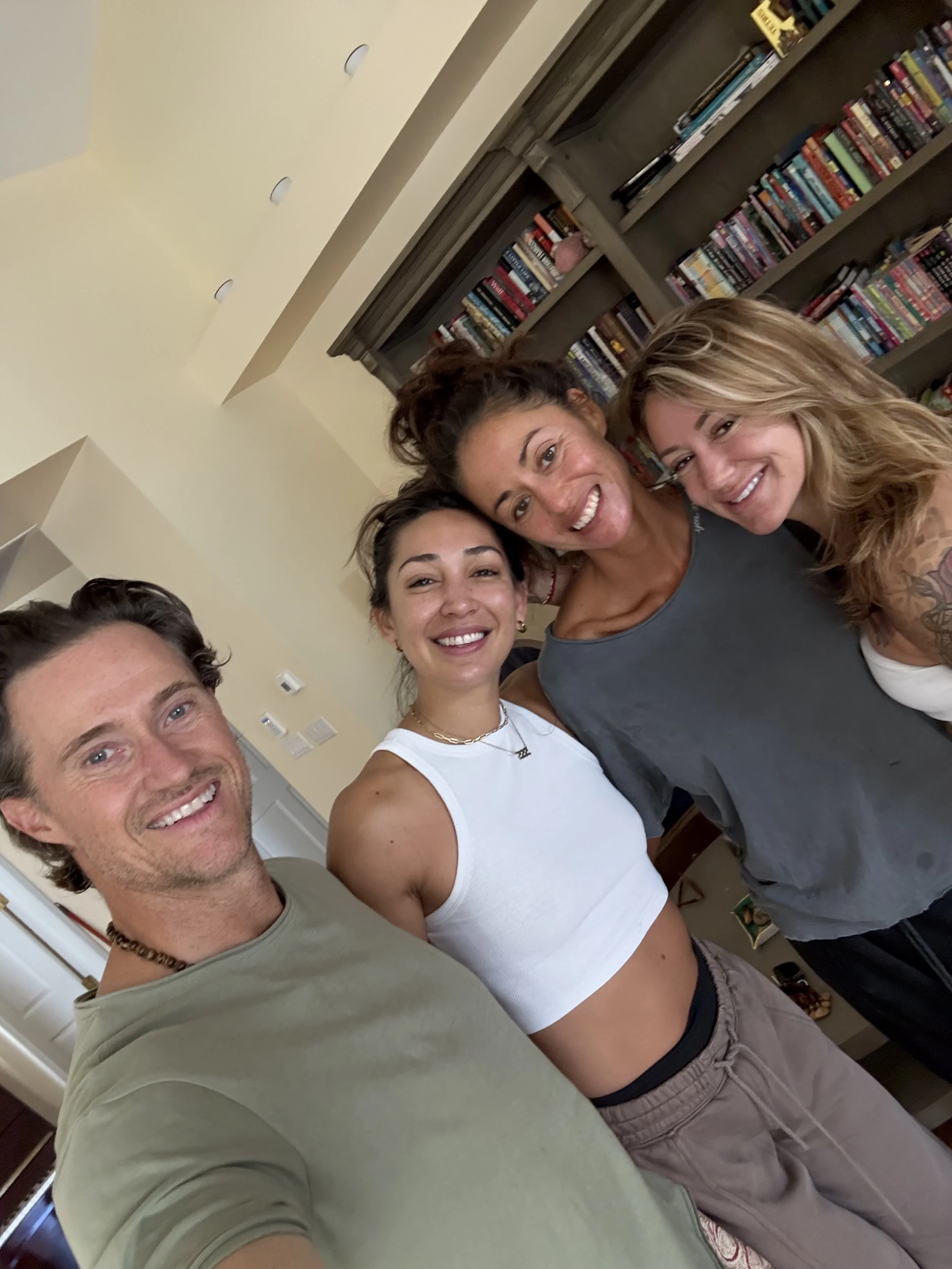 Four friends smiling and posing together in a room with a bookshelf in the background.