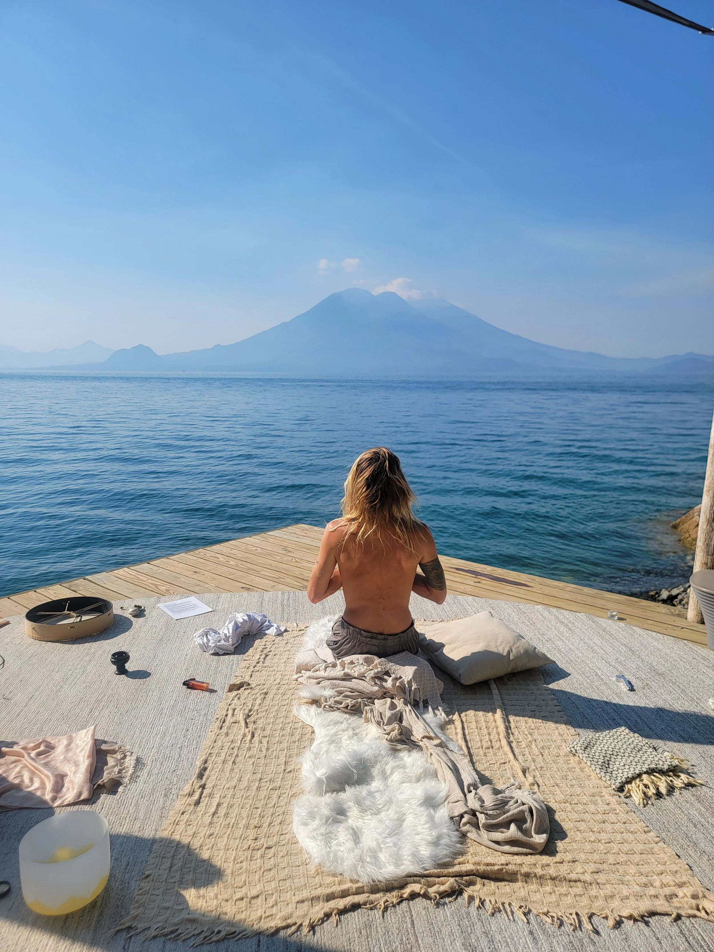 A person with long hair and a tattoo on their arm, sits cross-legged on a blanket on a dock, facing a large body of water with a mountain in the distance, under a clear blue sky.