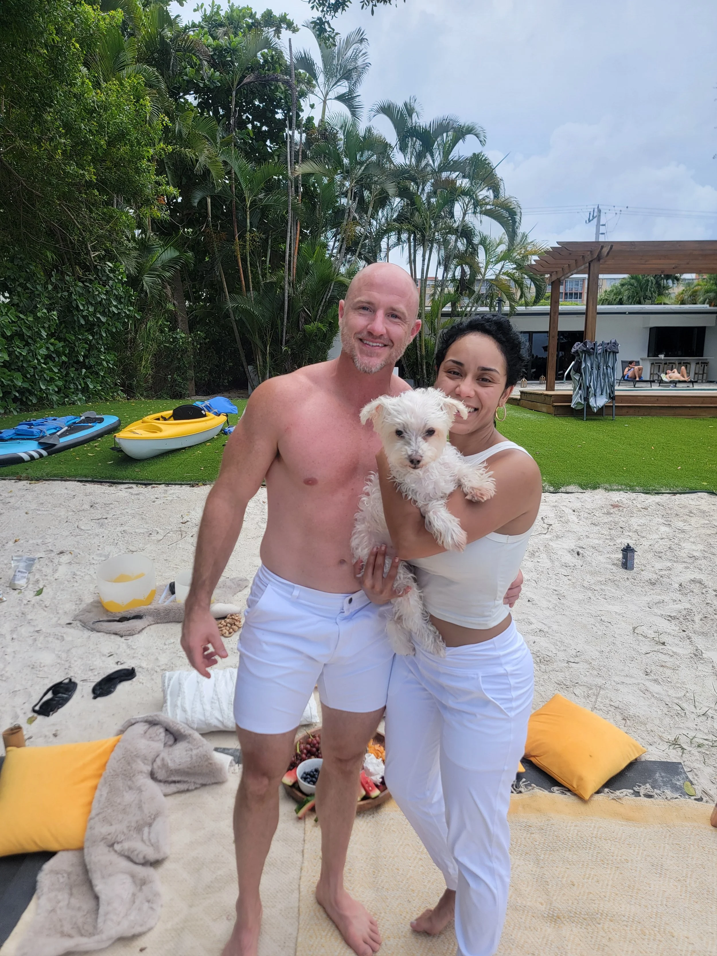 A smiling couple with a small white dog at the beach. The man is shirtless and wearing white shorts, and the woman is wearing a white tank top and white pants. They are standing on a sandy area with blankets and pillows. In the background, there are paddleboards, lush green trees, and a patio with people lounging.