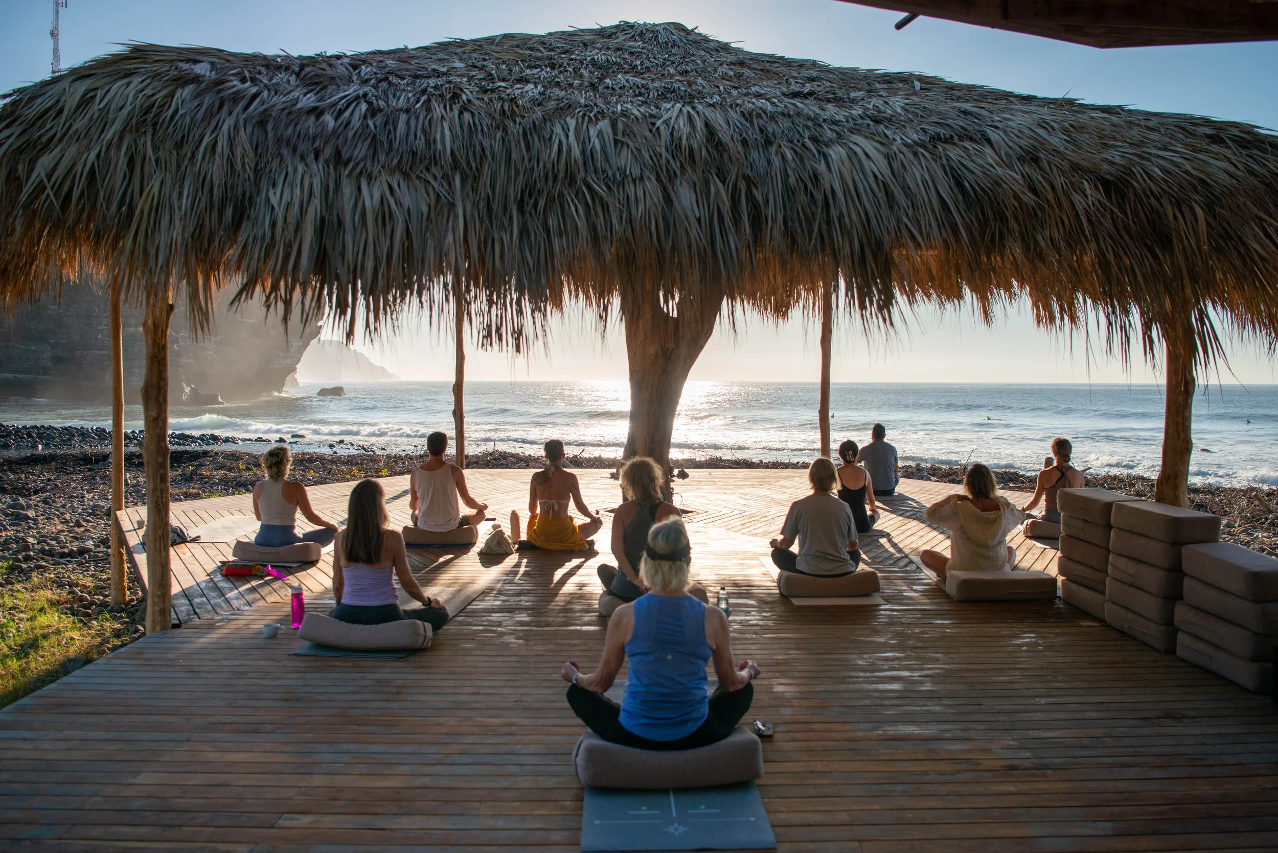 A group of people practicing yoga on cushions on a wooden deck under a thatched roof, facing the ocean at sunset.