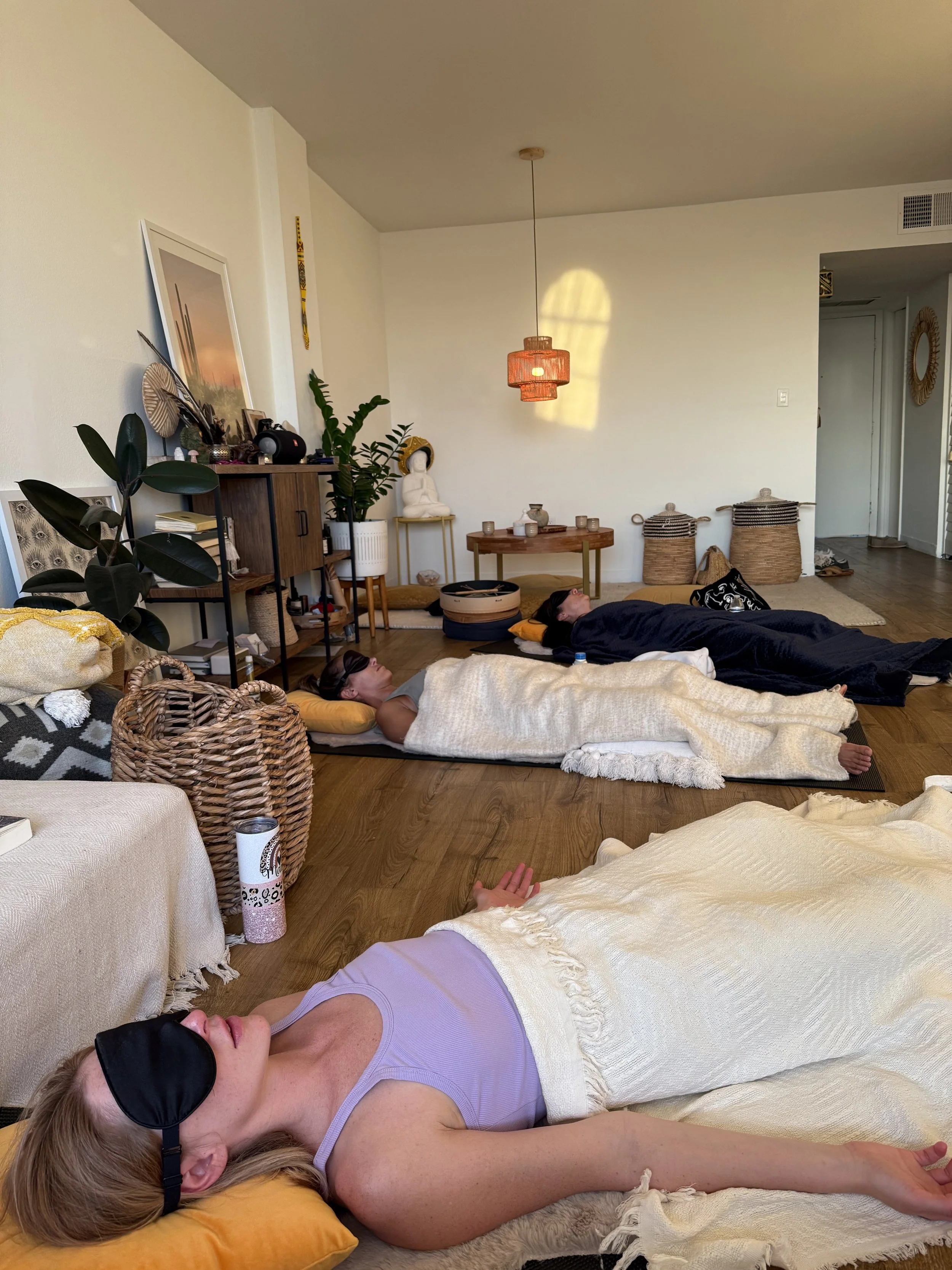 Three people lying on yoga mats doing breathwork, one with a sleep mask, resting in a bright, cozy living room with bohemian decor.