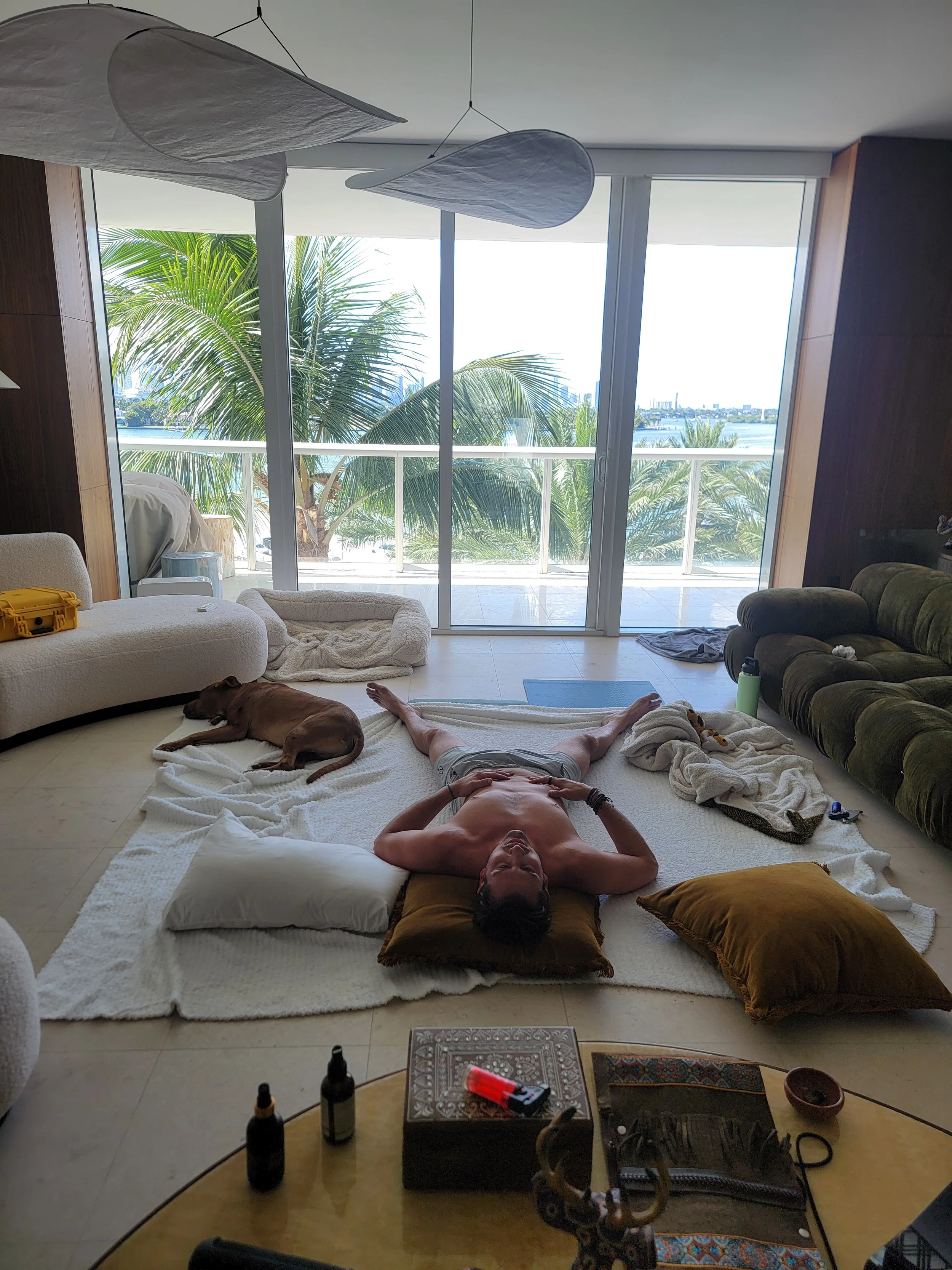 A man lying shirtless on a white blanket on the floor of a living room, with his hands on his stomach, surrounded by pillows. A dog is sleeping nearby. Large glass sliding doors leading to a balcony with palm trees and a body of water in the backgrou