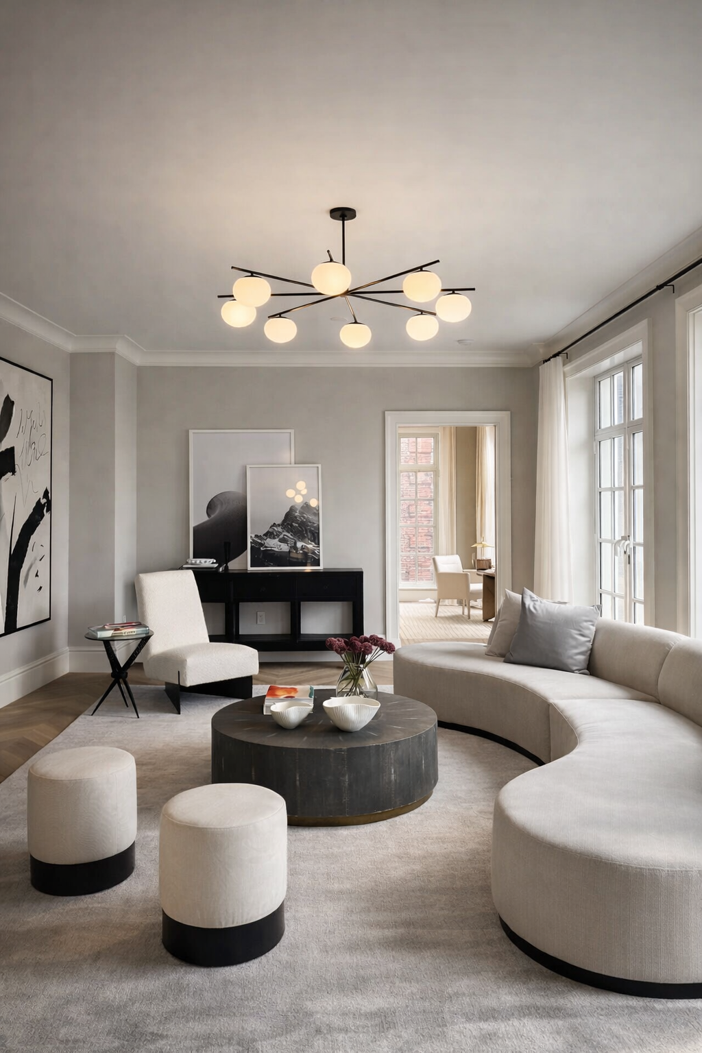 Modern living room with white curved sofa, two white ottomans, black round coffee table with decorative bowls and flowers, abstract wall art, and large windows letting in natural light.