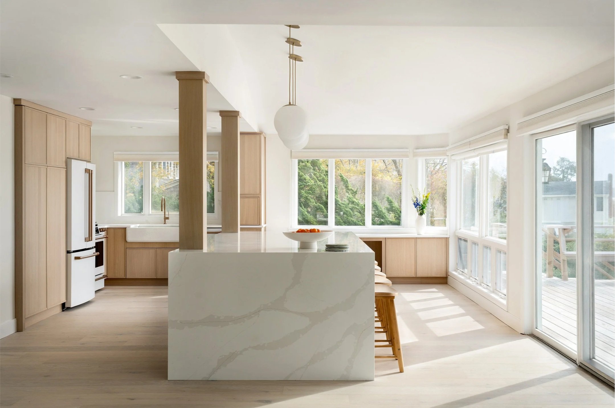 Bright and modern kitchen with large windows, light wood cabinets, a white marble island, and a dining area with wooden stools.