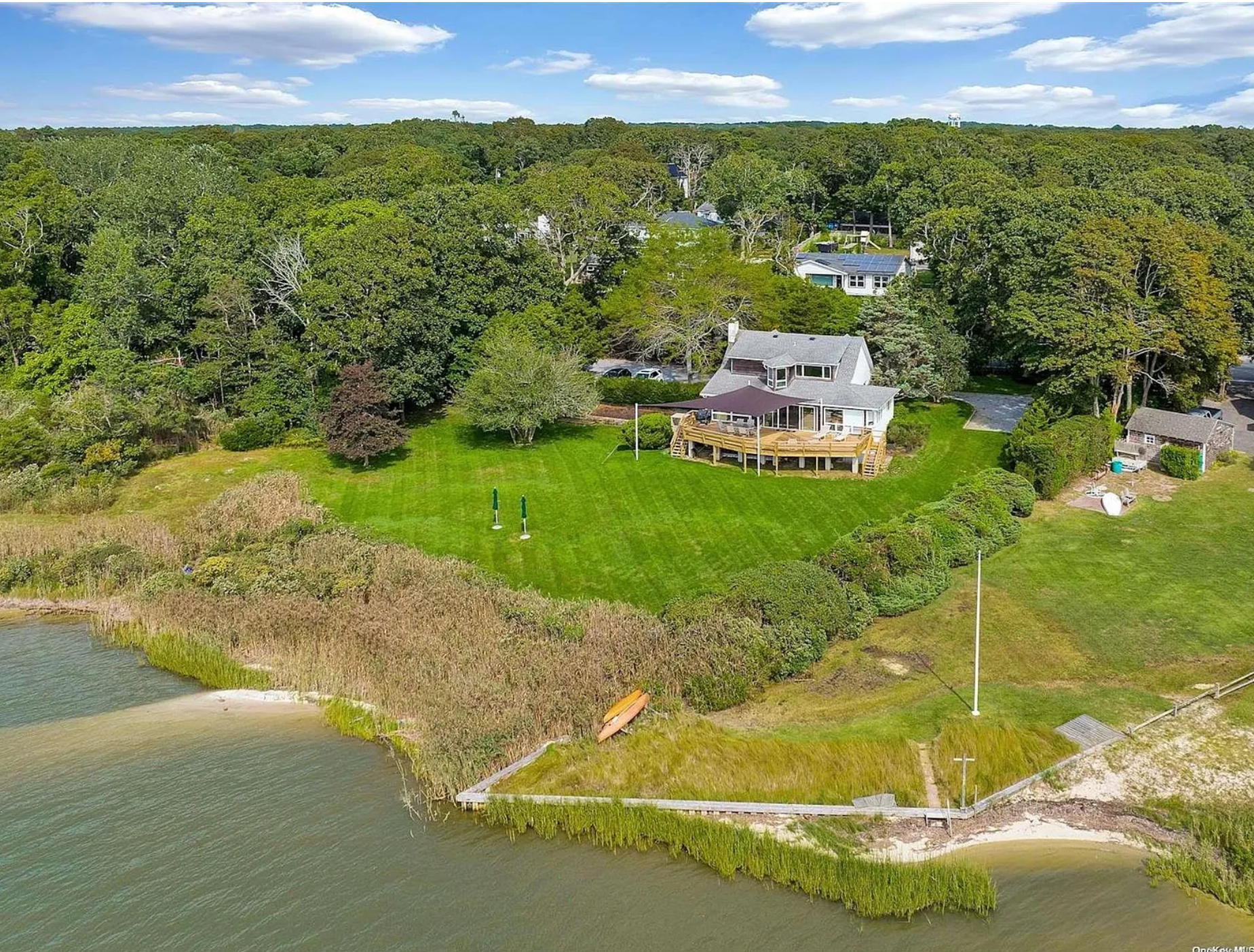 Aerial view of a house with a deck, lush green lawn, trees, and a waterfront shoreline