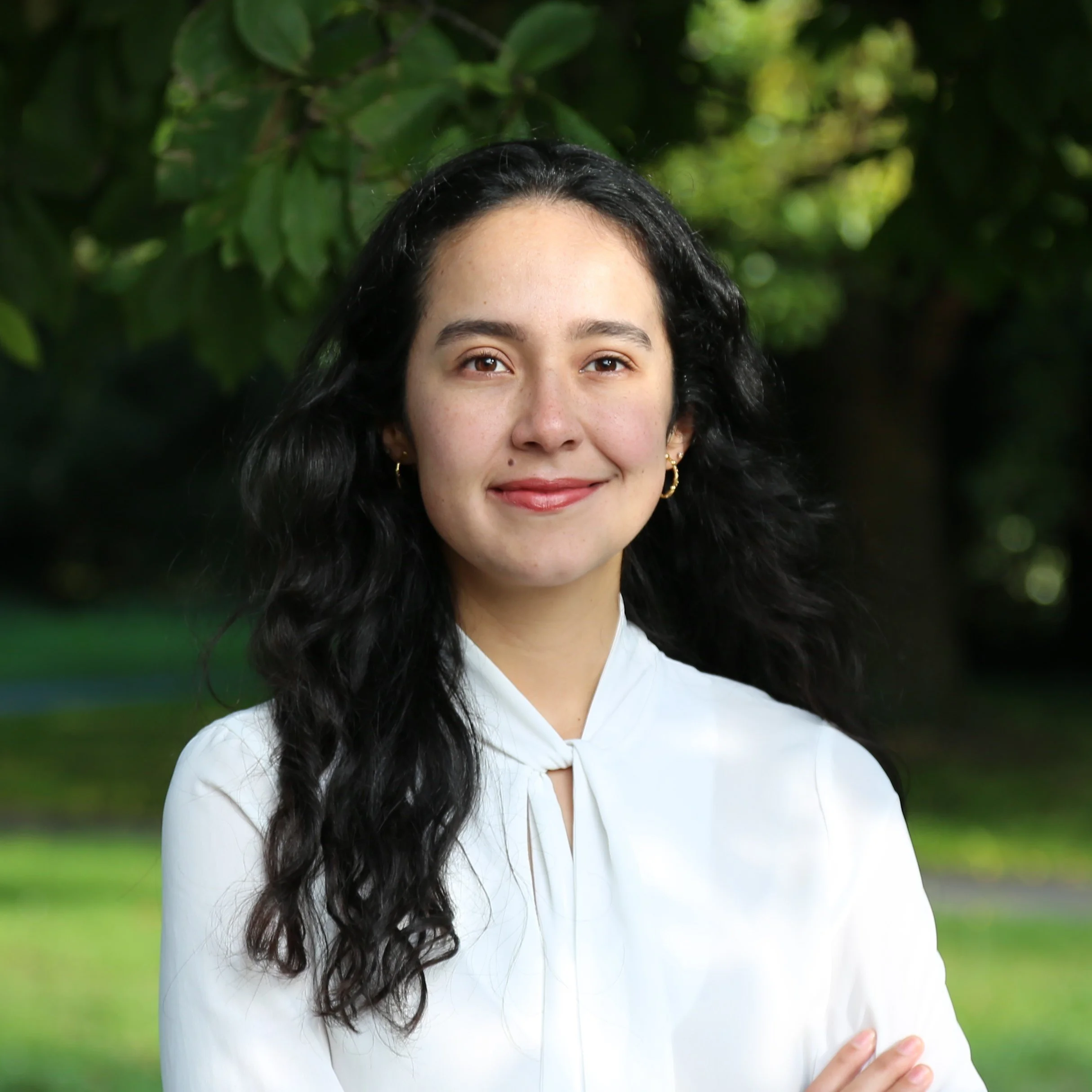 Portrait of a young woman with long, wavy black hair wearing a white blouse, standing outdoors with greenery in the background.