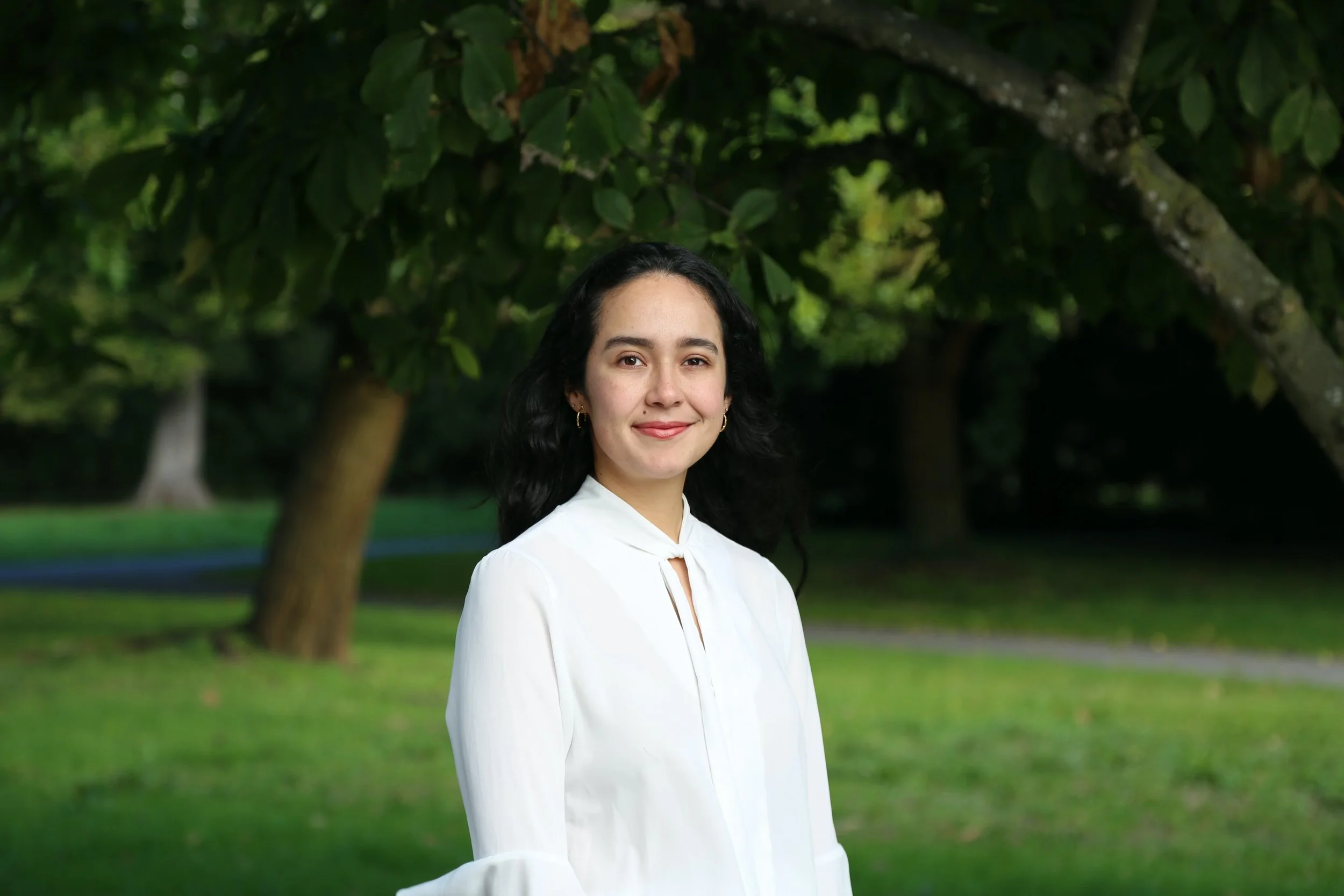 A young woman with dark, wavy hair and earrings, wearing a white blouse, stands outdoors with trees and grass in the background.