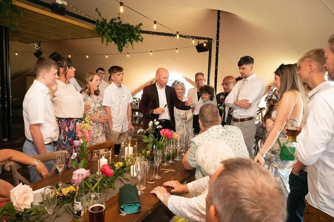 Group of people gathered around a table at a celebration, with a man speaking into a microphone. The table has flowers and candles, and the setting appears to be a decorated party or wedding reception inside a tent.