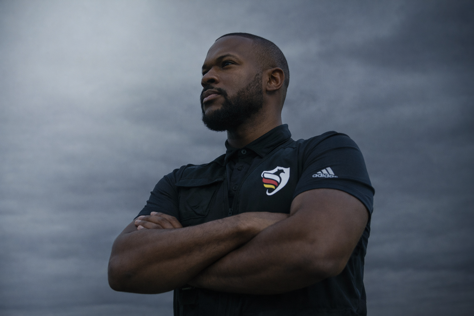 Mann steht mit verschränkten Armen vor dunklem Himmel, trägt schwarzes Adidas-Poloshirt mit logos und Blick in die Ferne nach rechts.