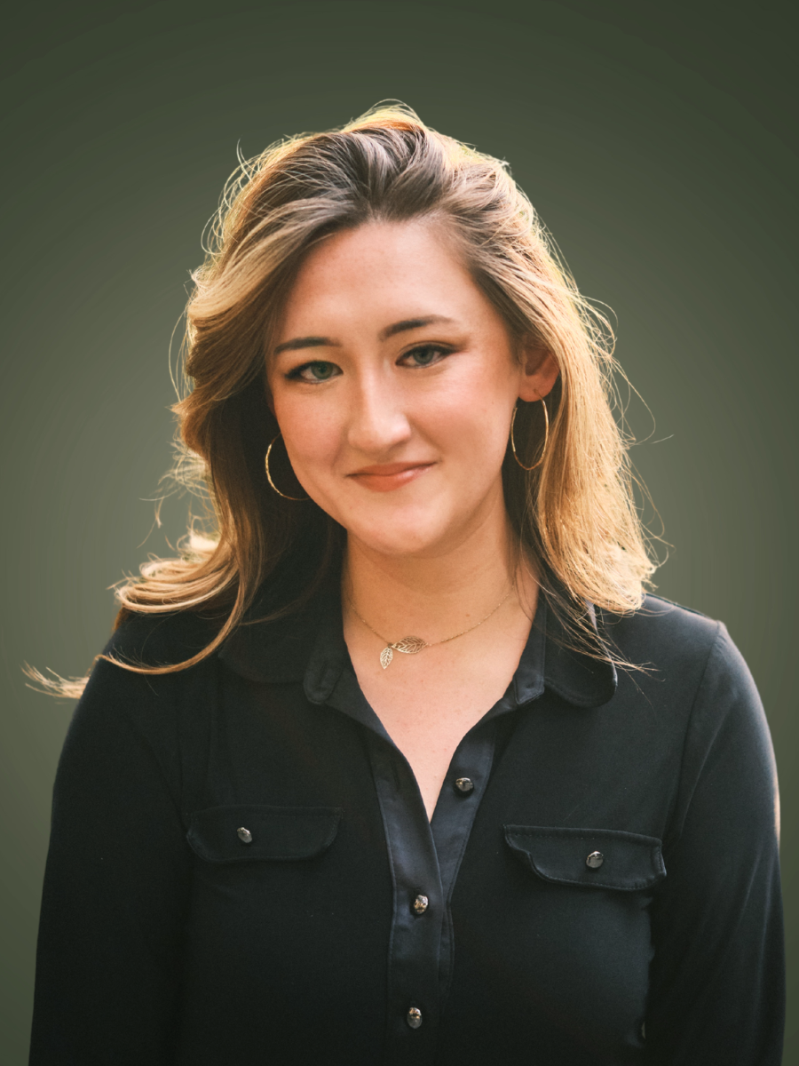 A young woman with blonde hair wearing hoop earrings and a black button-up shirt, standing against a blurred green background.