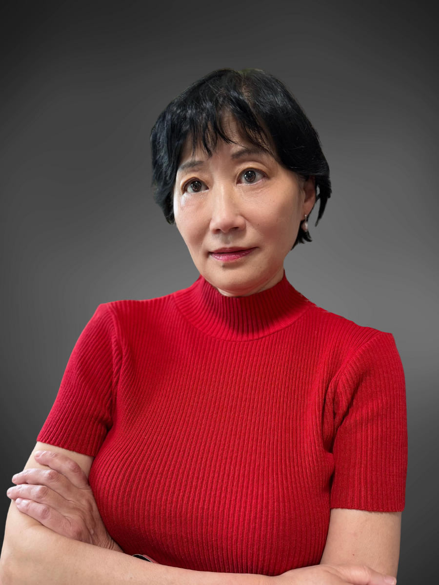 A woman with short black hair wearing a red ribbed turtleneck sweater, crossing her arms against a dark gray background.