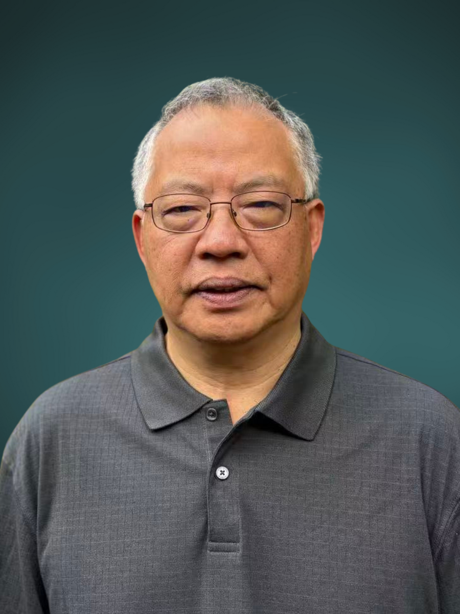 A middle-aged Asian man wearing glasses and a dark gray polo shirt looking at the camera with a neutral expression, standing against a plain dark green background.