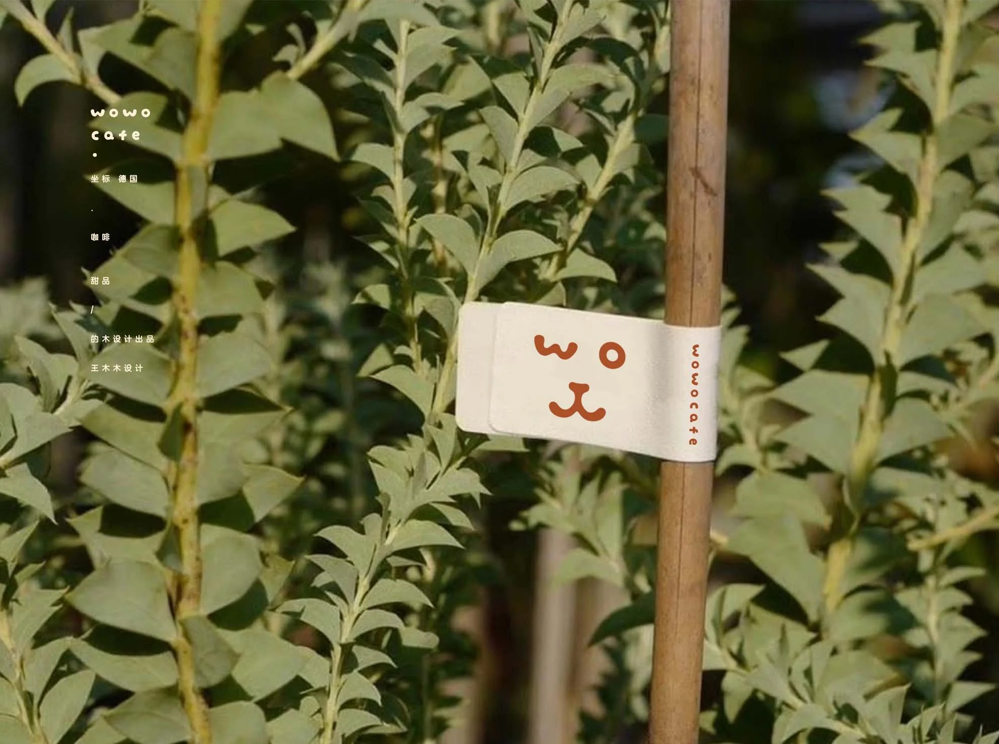 A wooden stake with a white tag attached, which has a simple drawing of an animal face and the word 'woocafe' written vertically. The stake is among green, leafy plants with sunlight filtering through.