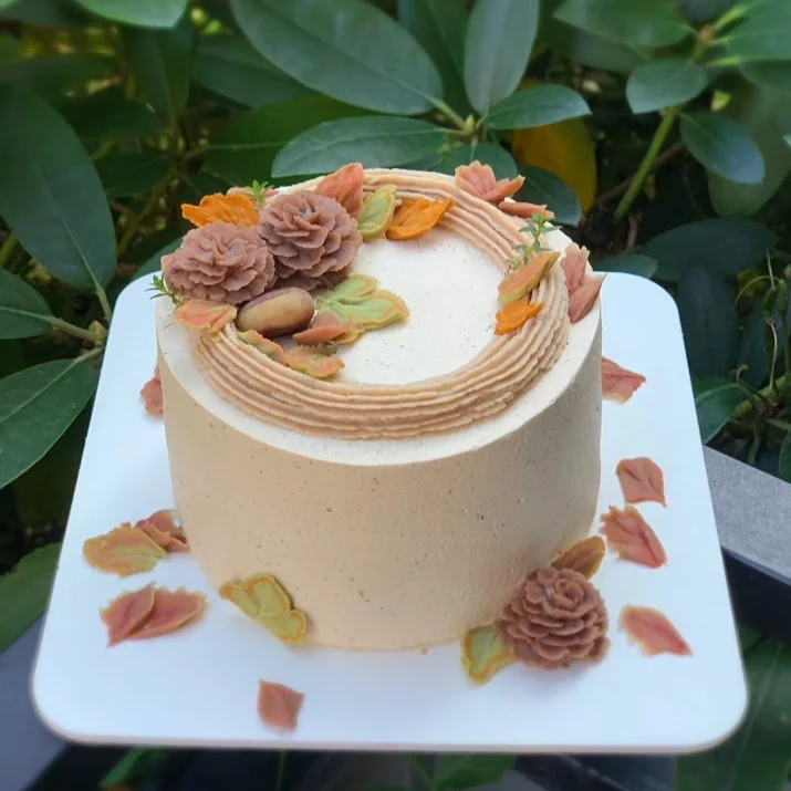A round, white frosted birthday cake decorated with peach and green floral designs and placed on a white square plate, with a background of green leaves.