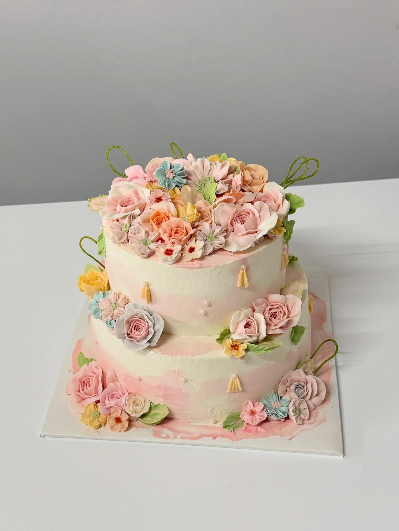 Two-tiered wedding cake decorated with pastel-colored roses, flowers, pearls, and tassels, topped with a floral arrangement.