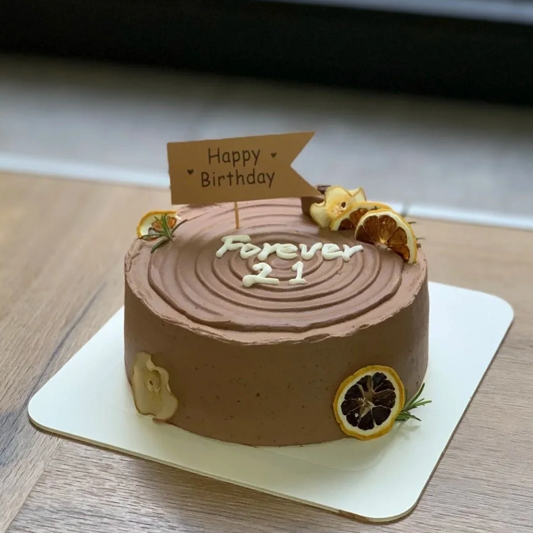 Chocolate birthday cake decorated with dried lemon slices, a small yellow fondant duck, and a 'Happy Birthday' flag, with 'Forever 21' written in white icing on top.