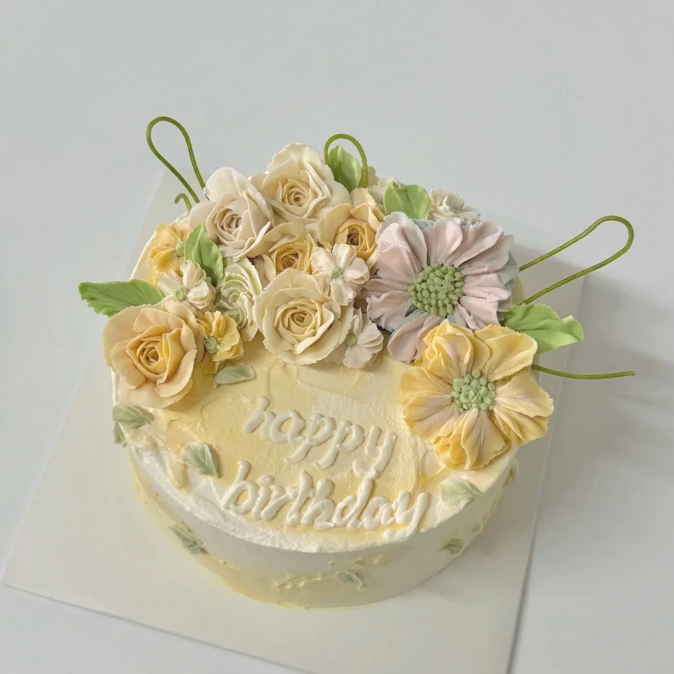 A round birthday cake decorated with ivory roses, pink daisies, green leafy accents, and the words 'happy birthday' written in white icing on top.