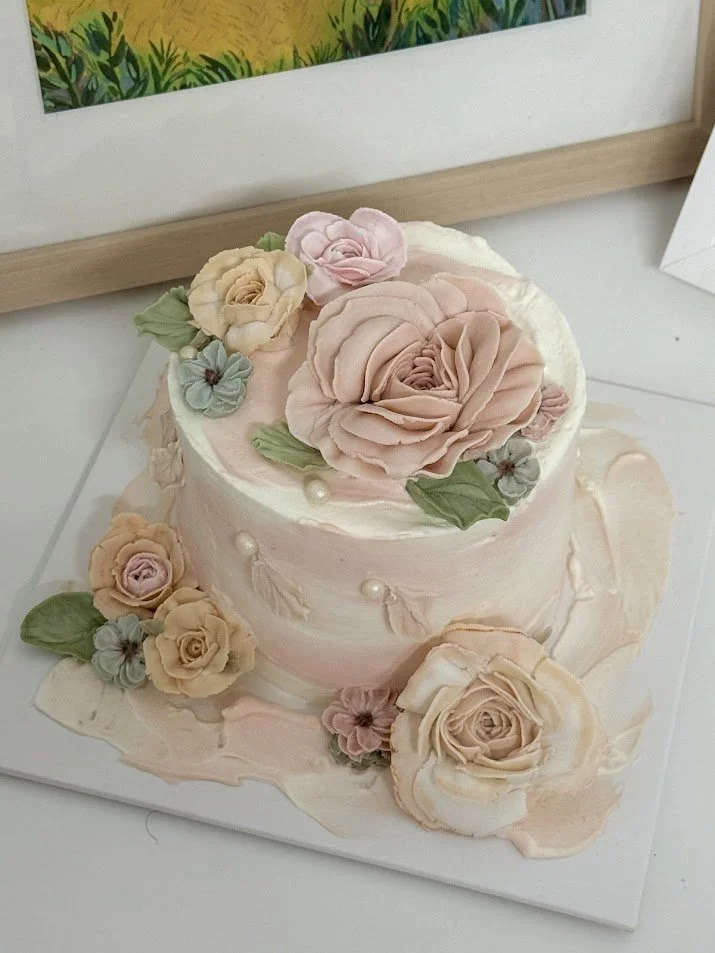 A white and pink decorated cake with large pearl accents, topped with pink and yellow roses, and additional roses and leaves around the base, placed on a white surface.