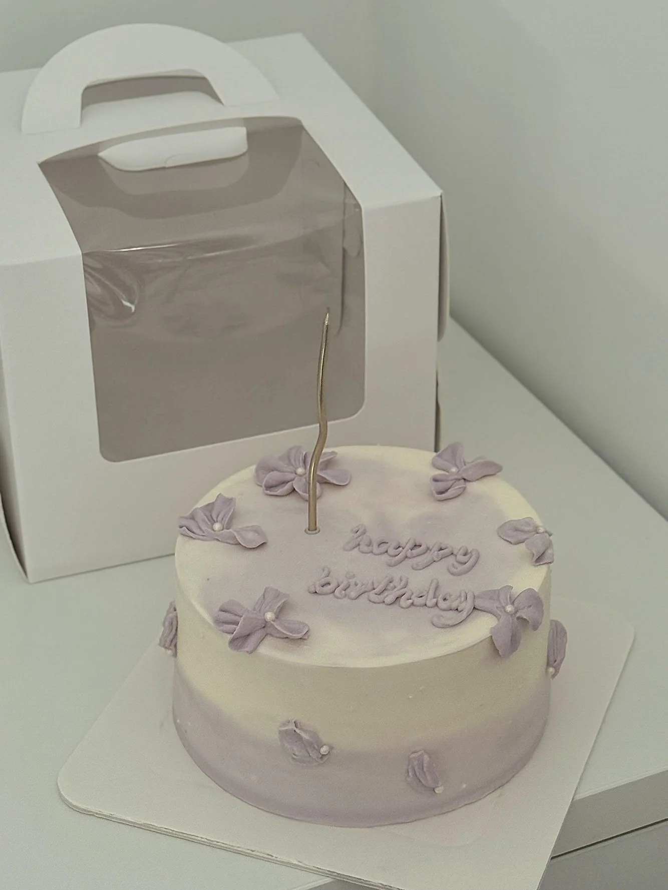 A round birthday cake with light purple and white frosting, decorated with purple bows and small white pearls, with a gold candle on top. The cake has a message that reads 'happy birthday' in purple icing. A white gift box with a window and handle is in the background.