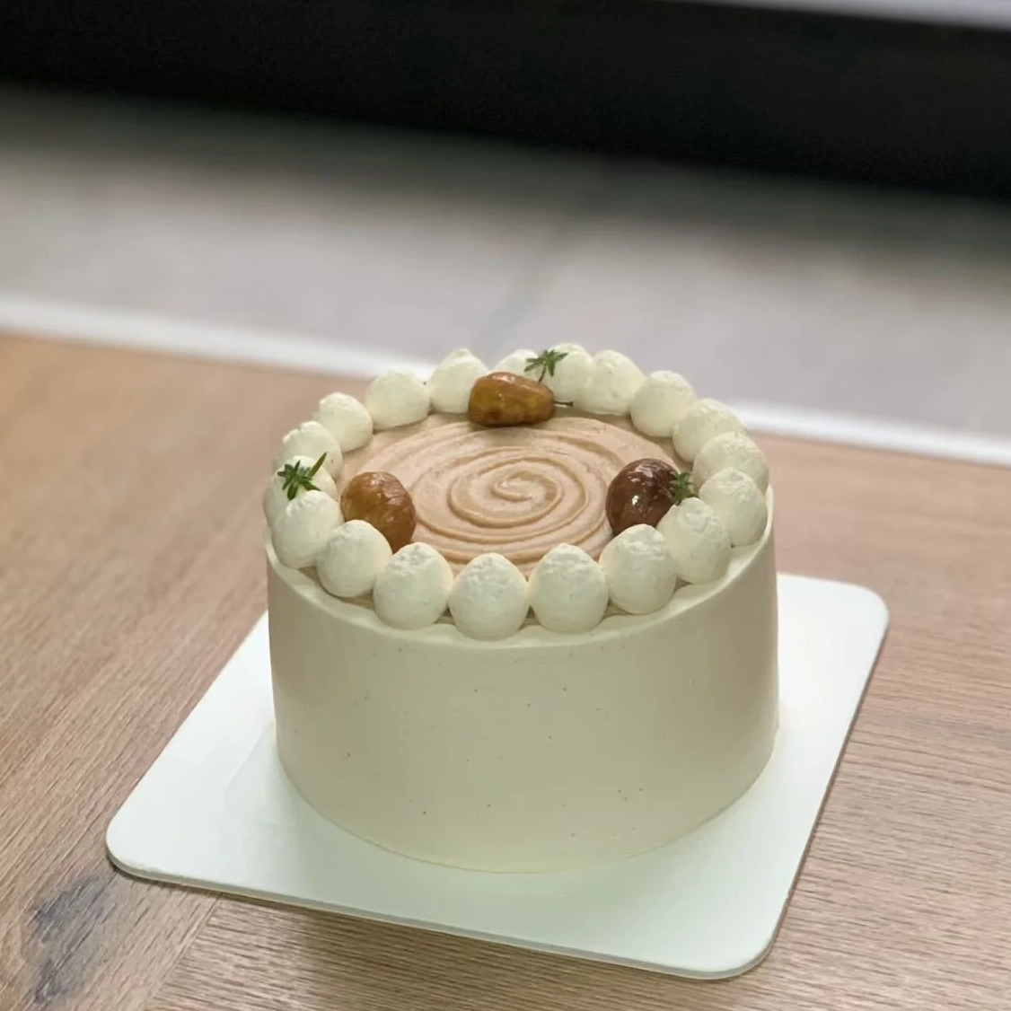 Round cake with white frosting, decorated with white dollops of frosting around the top edge, brown caramelized pieces, and small green herbs on top, sitting on a white square base on a wooden surface.