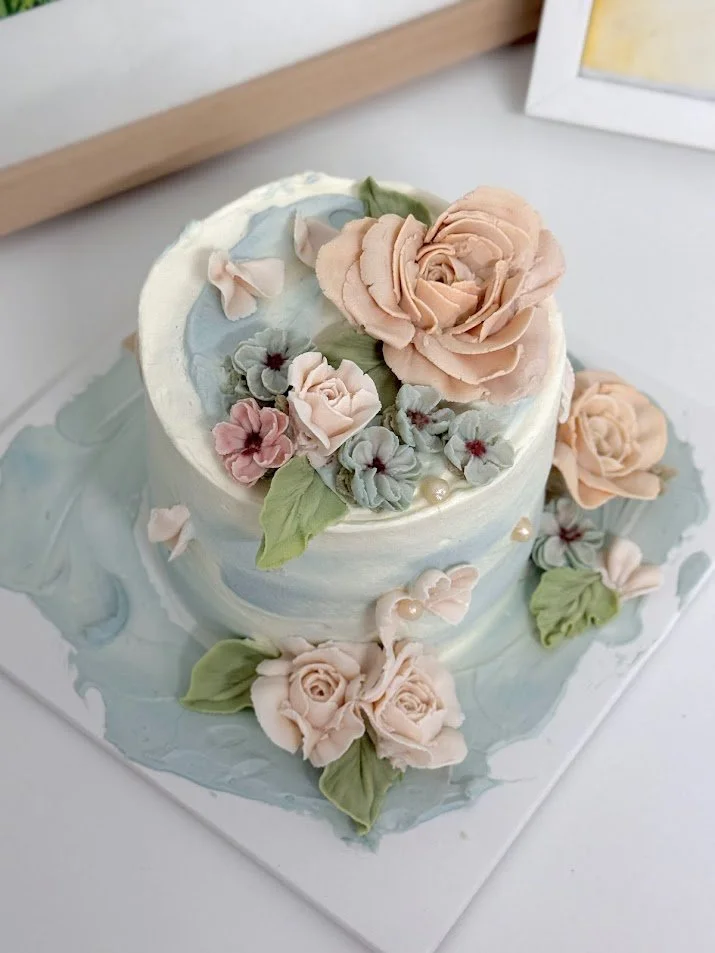 A two-tiered cake decorated with pastel-colored buttercream flowers, including roses, and green leaves, on a marble-like cake board.