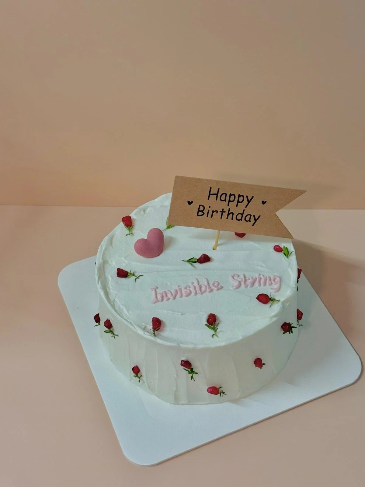 A white birthday cake decorated with small red berries and a pink heart-shaped decoration. A brown paper flag reads 'Happy Birthday' and pink icing spelling says 'Invisible String' on top of the cake, which is on a white square board on a pale pink surface.