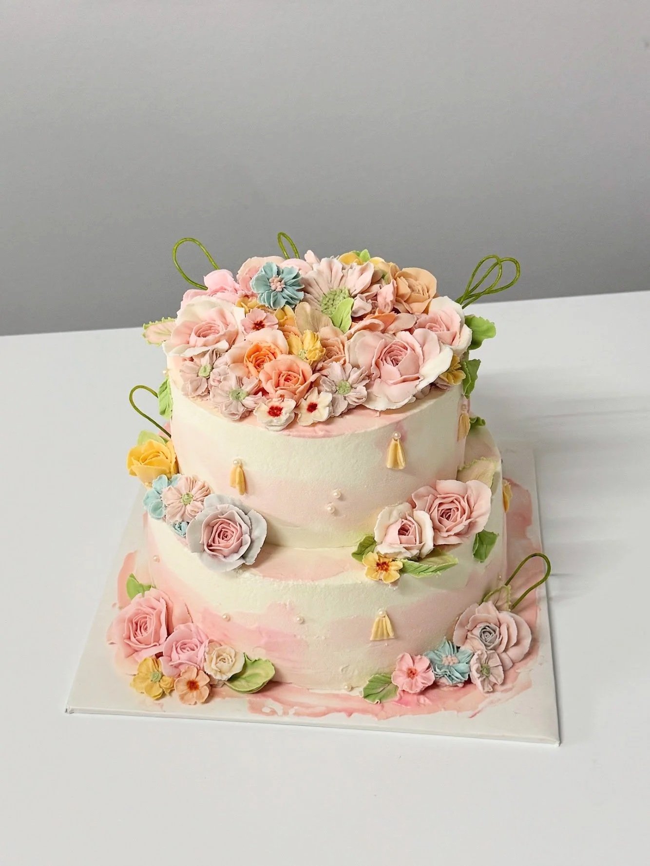 Blumencake. Zweistöckige Hochzeitstorte mit zartfarbenen Rosen, Blüten und grünen Blättern verziert, gekrönt von einer Vielzahl von Fondantblumen und filigranen Akzenten – präsentiert auf einer weißen Fläche.