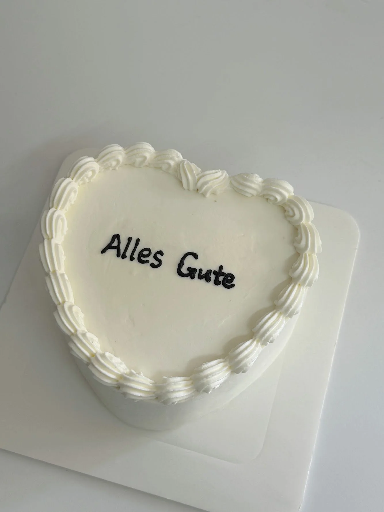 White heart-shaped cake with piped white frosting border and the words "Alles Gute" written in black icing on top.