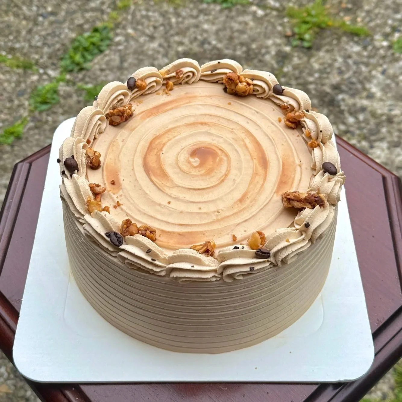 Round cake with light brown frosting, decorated with piped grayish frosting border and topped with caramel sauce, walnuts, and chocolate chips.