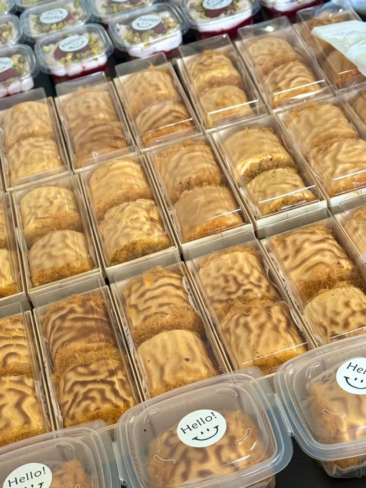 Rows of individually packaged cookies with icing drizzle in clear plastic containers, some with 'Hello!' stickers with smiley faces on top.