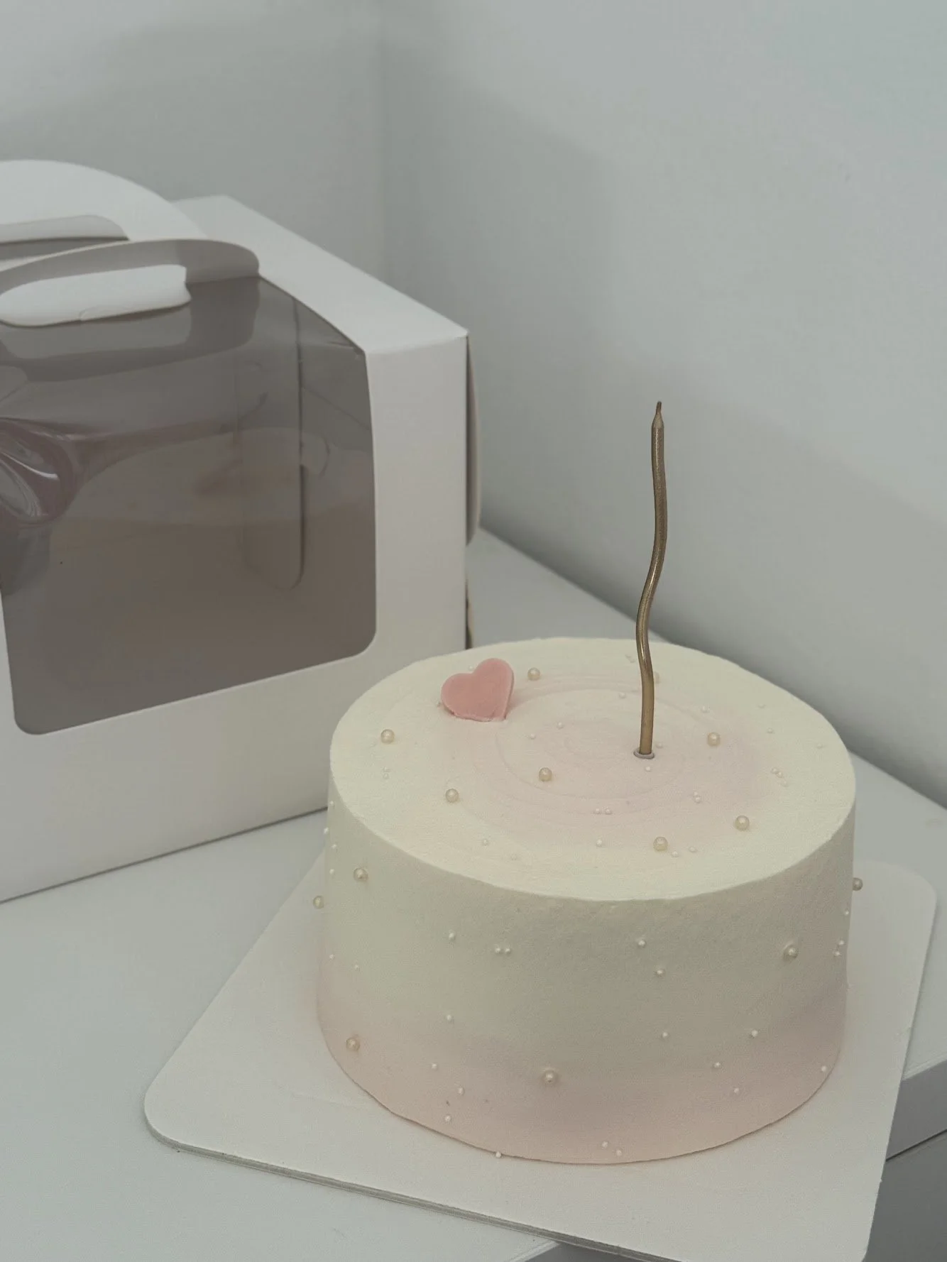 A white birthday cake with small pearl-like decorations, a pink heart-shaped decoration, and a metallic candle on top, placed on a white surface near a white box with a transparent window.