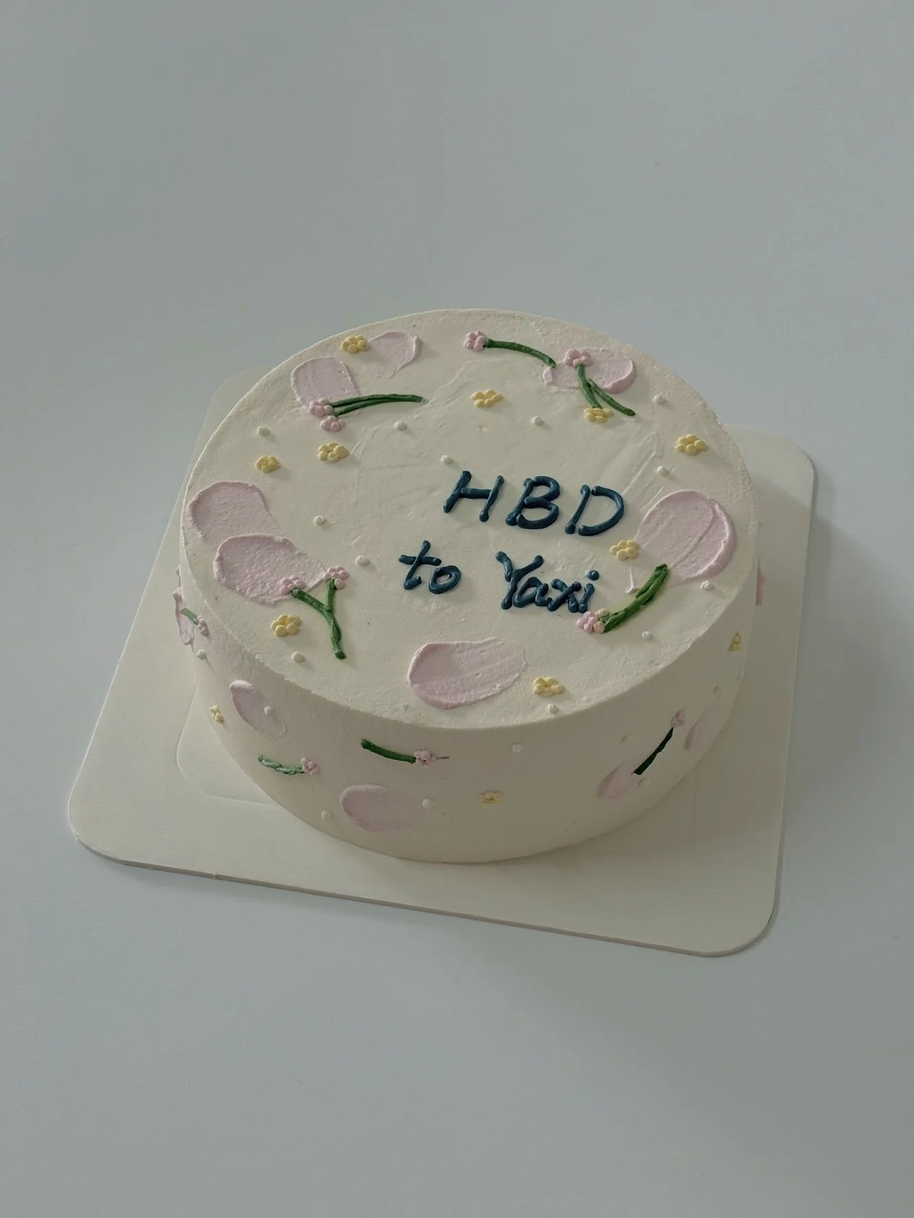 A round white cake decorated with pink and yellow icing details and green decorations, with the message "HBD to Yayi" written in blue icing on top.