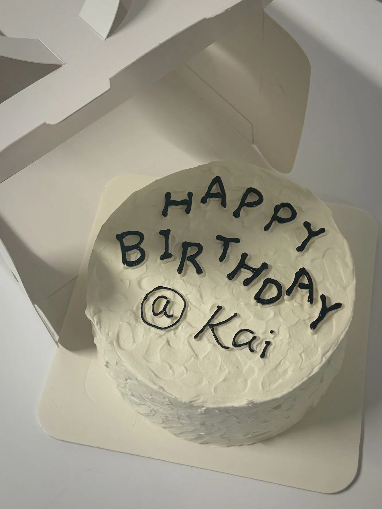 White birthday cake with black writing that says 'HAPPY BIRTHDAY @ Kai'. The cake is in a white box.