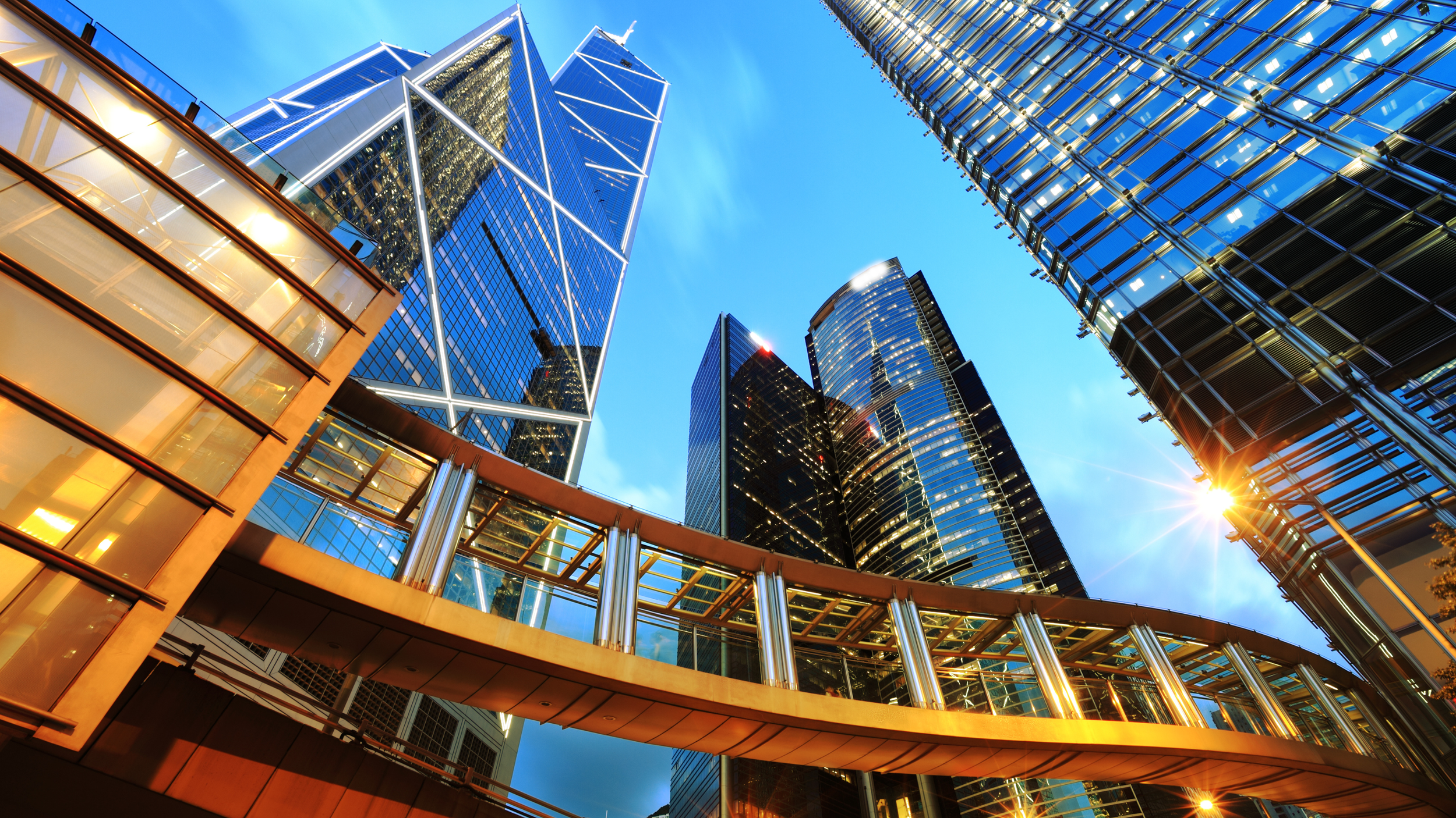 Looking up at modern glass skyscrapers in a city at sunset with a pedestrian bridge in foreground