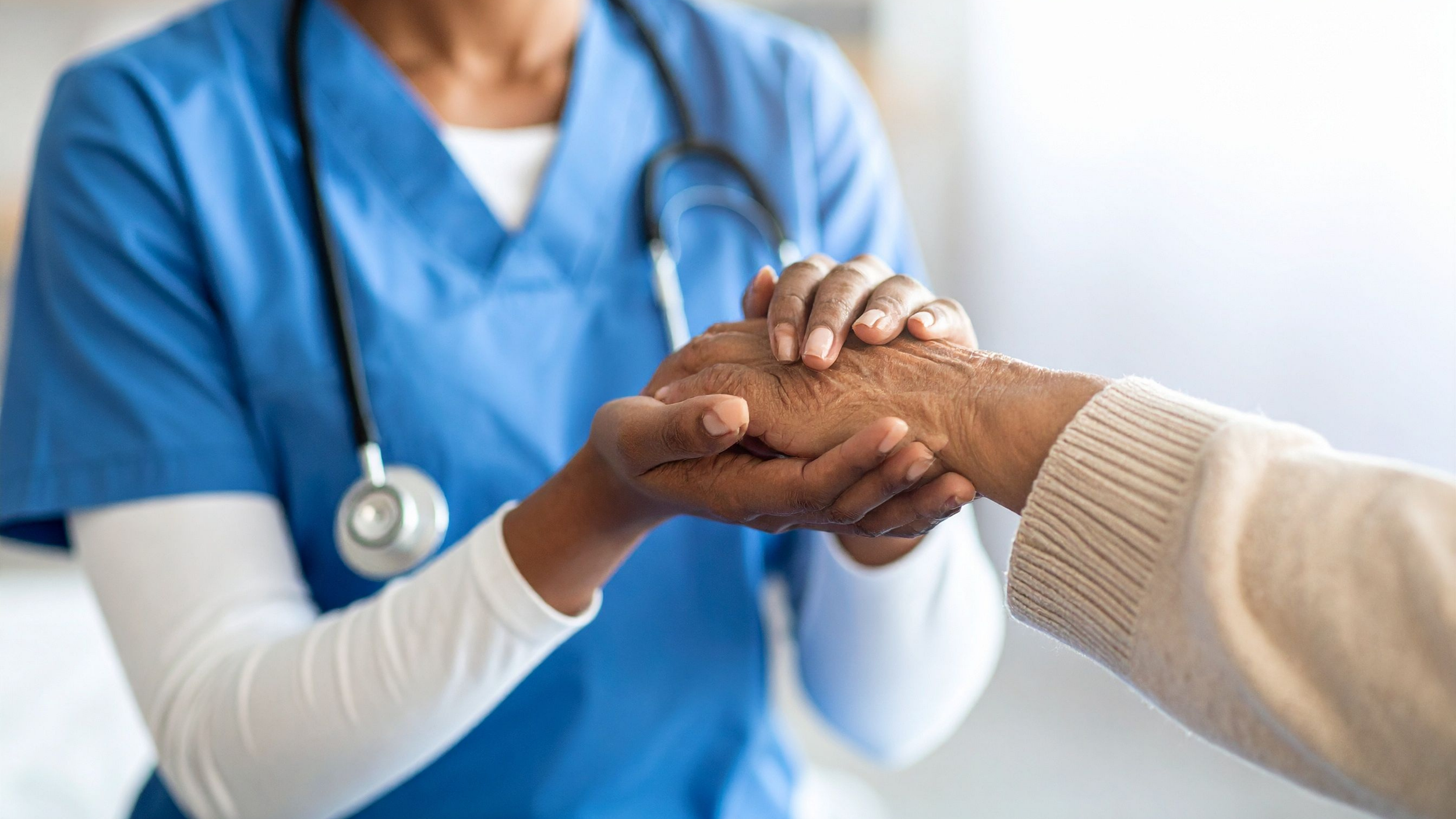 A healthcare worker in blue scrubs and a stethoscope holding and comforting an elderly person's hand.