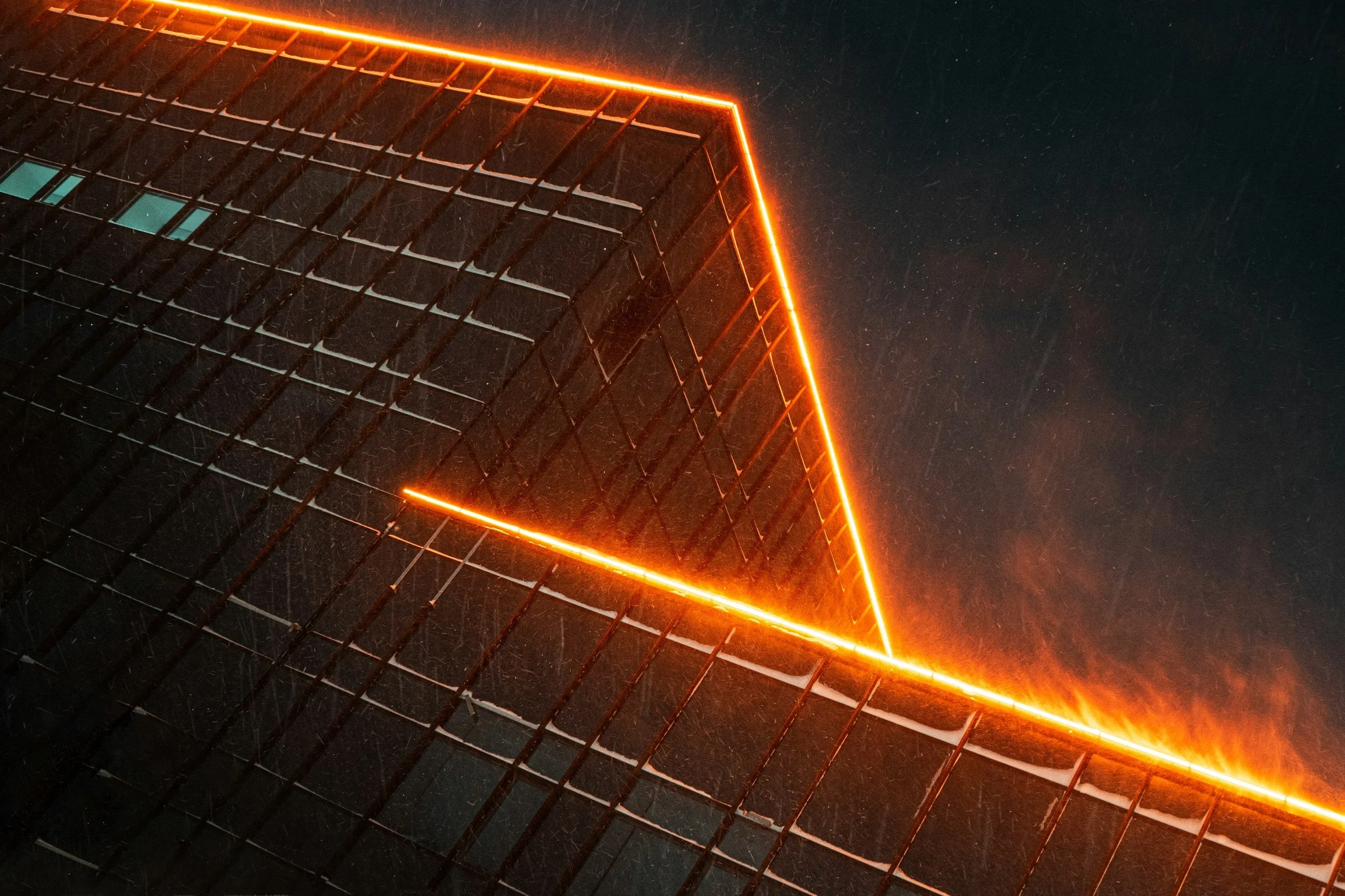 A close-up of a modern building with access to a black sky showing a red neon outline along the top edges of the building and the snow falling.