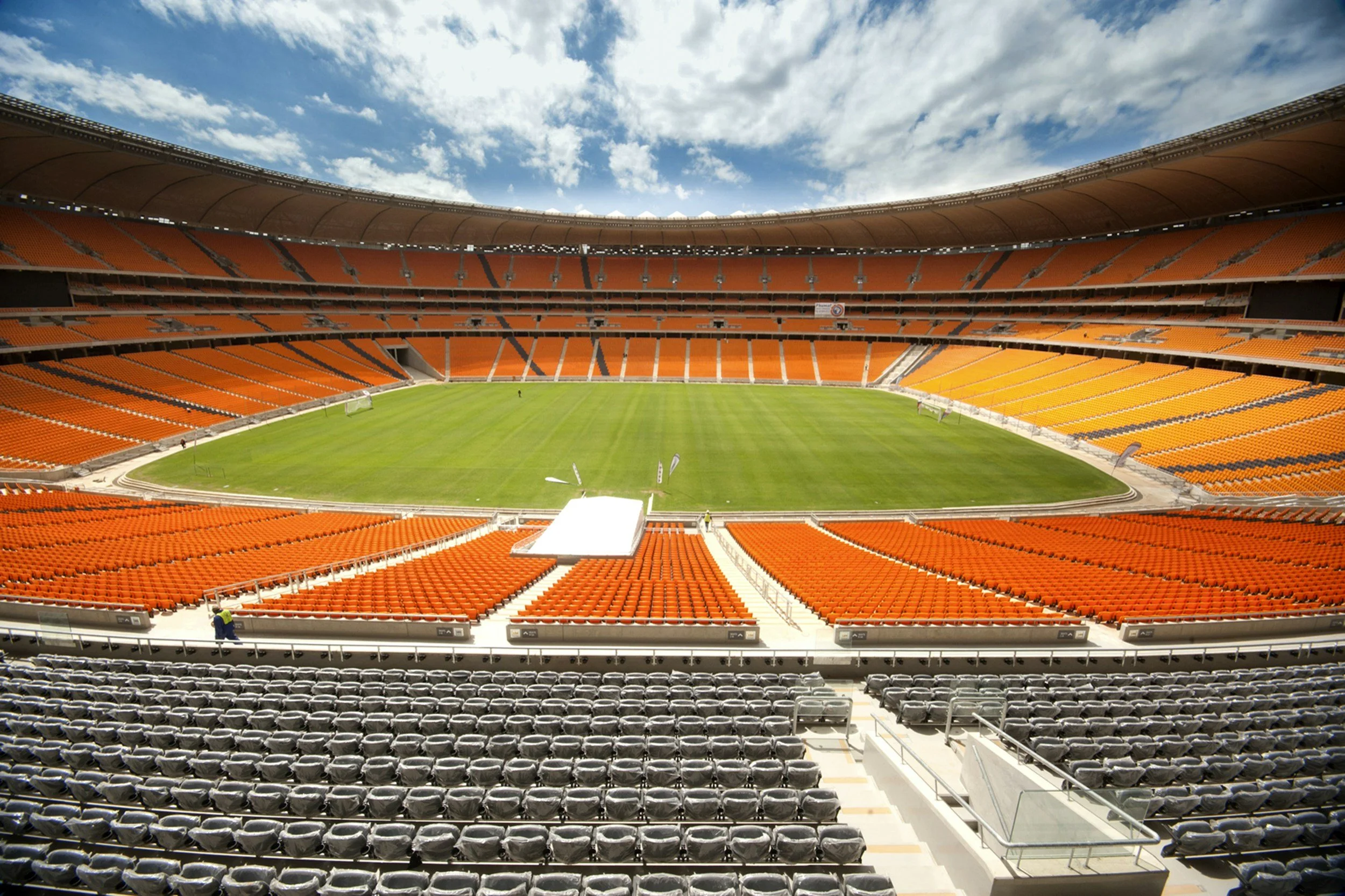Empty sports stadium with orange and black seats, green field, blue sky with clouds.