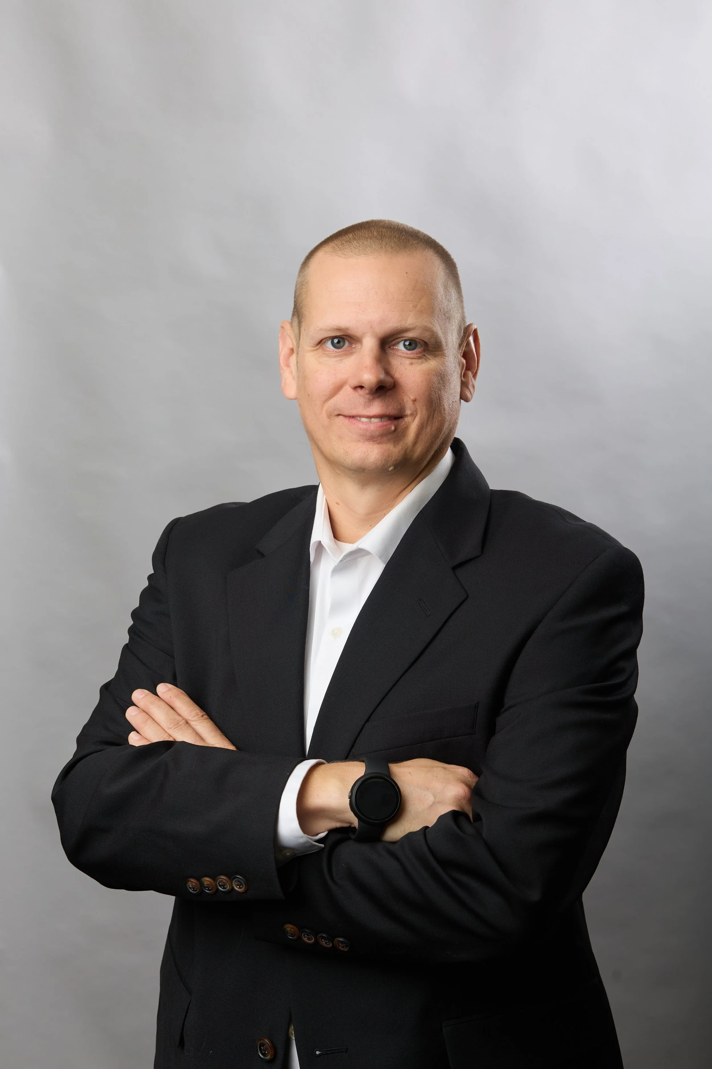 A man in a black suit with crossed arms, wearing a white dress shirt and a black smartwatch, posing against a gray background.