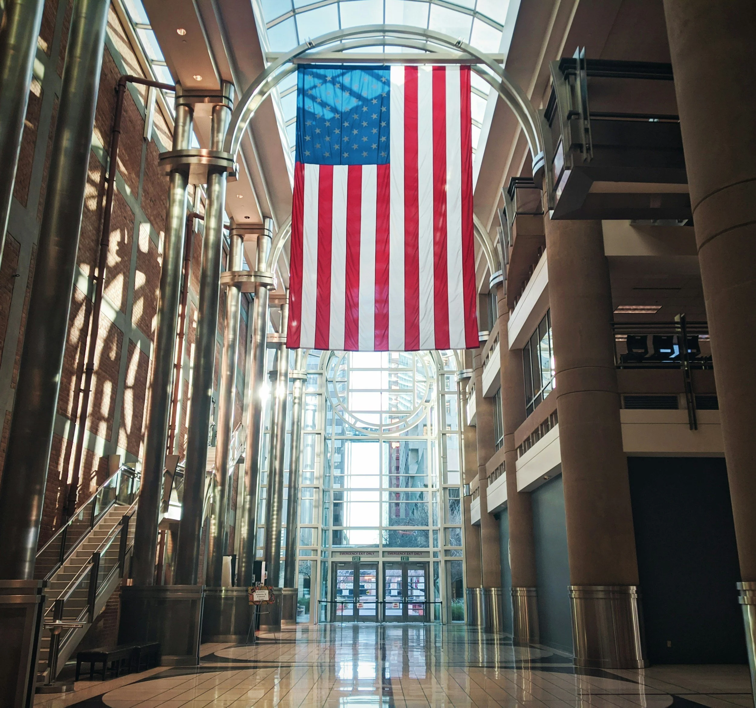 Interior of a modern building with a large American flag hanging from the ceiling, glass walls, and metal pillars.
