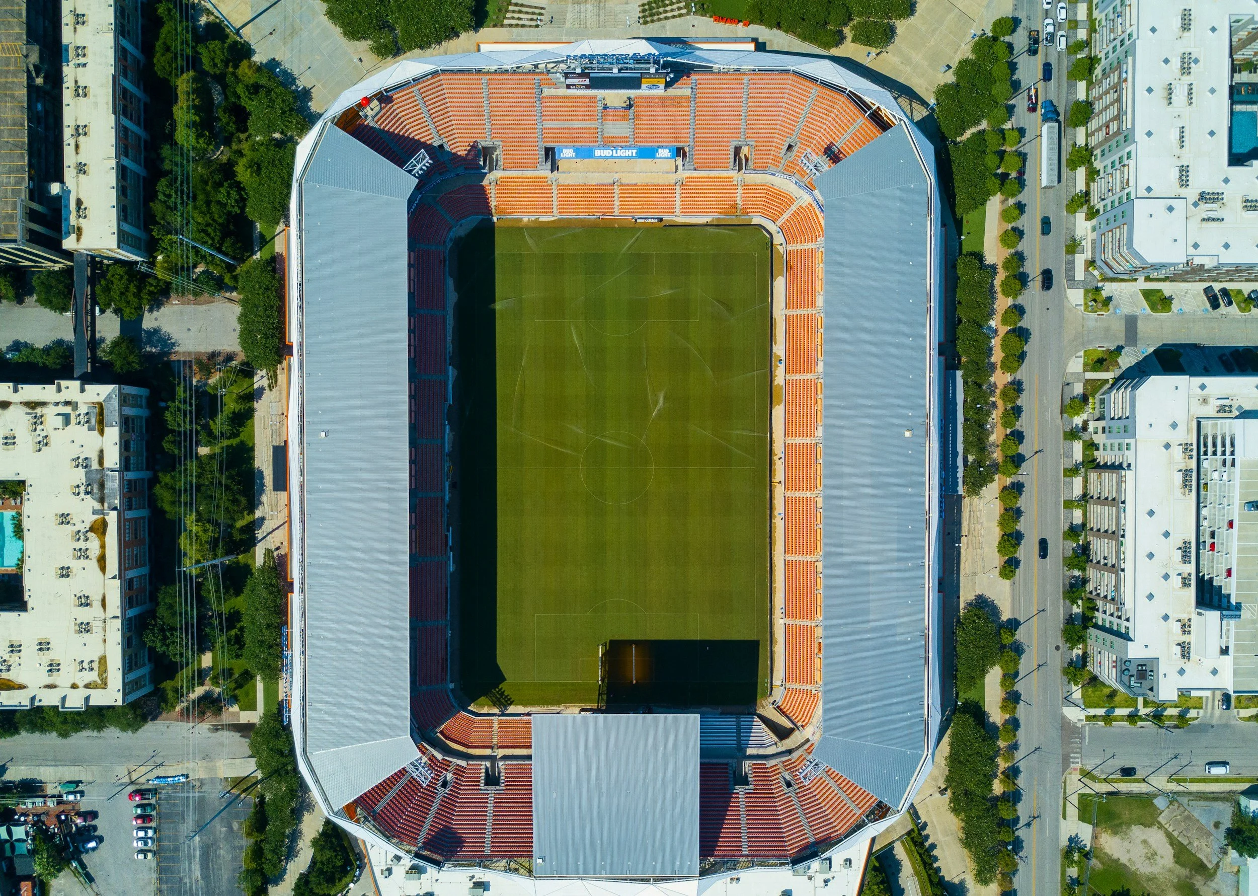 Bird's eye view of an empty football stadium with orange seating, surrounded by trees, buildings, and nearby streets.