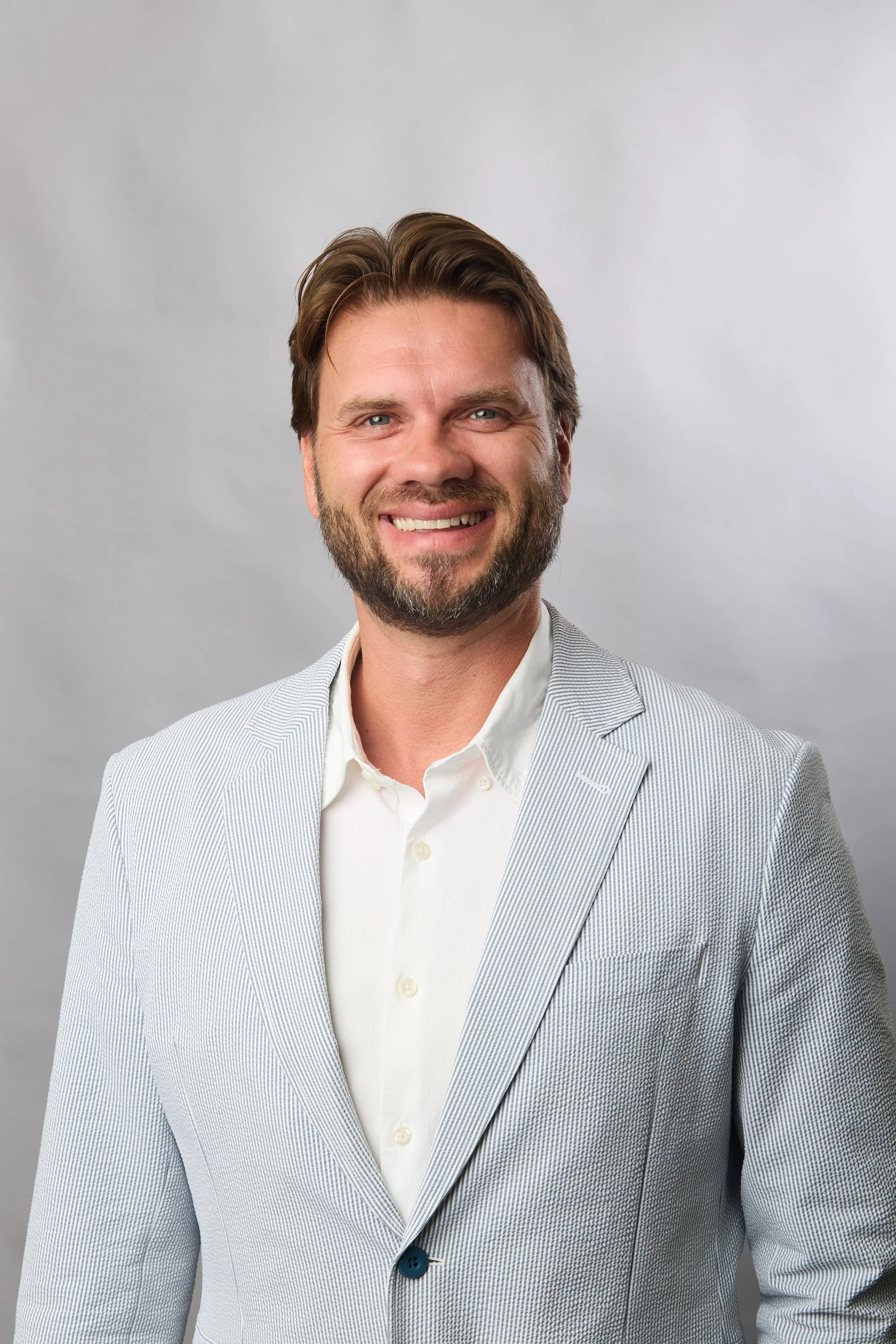 A smiling man with brown hair, beard, wearing a white dress shirt and light gray blazer, standing against a plain light gray background.
