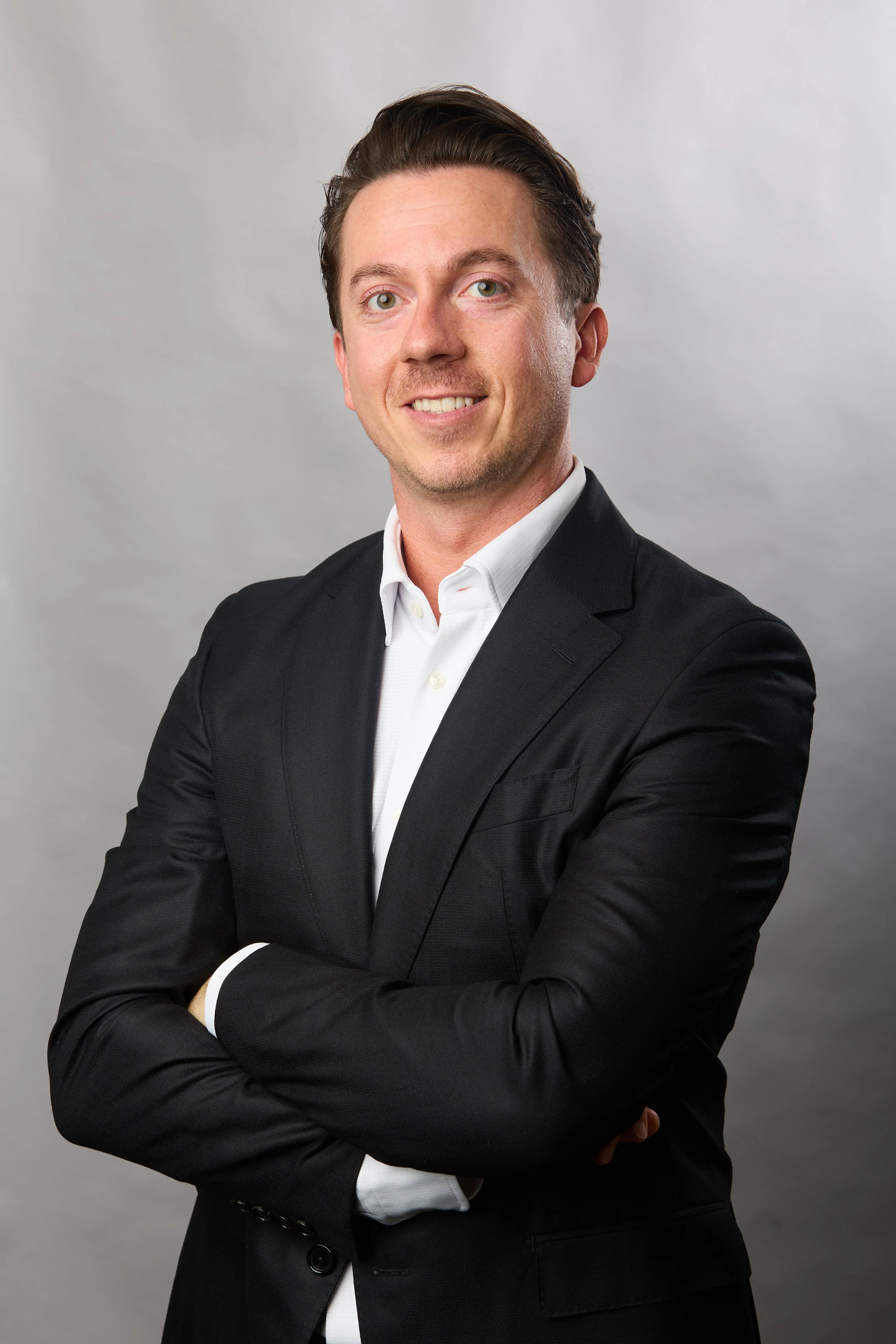 A young man with brown hair and fair skin, wearing a black suit jacket and white shirt, smiling with arms crossed in a professional headshot against a gray background.