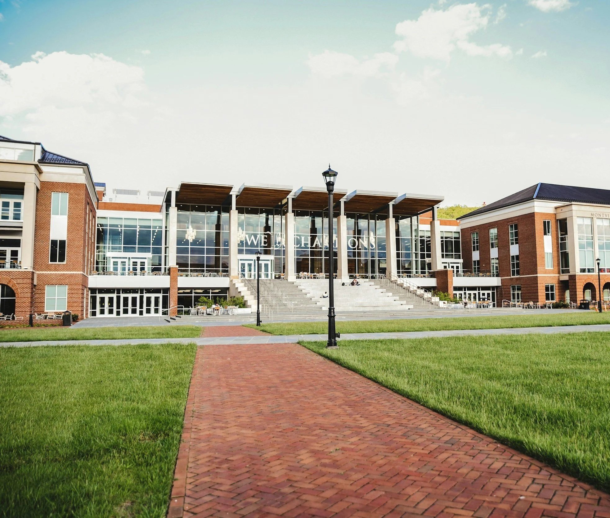 Exterior of a modern building with large glass windows and brick walls, located on a green lawn with a brick pathway leading up to the entrance, and a lamppost in the foreground.