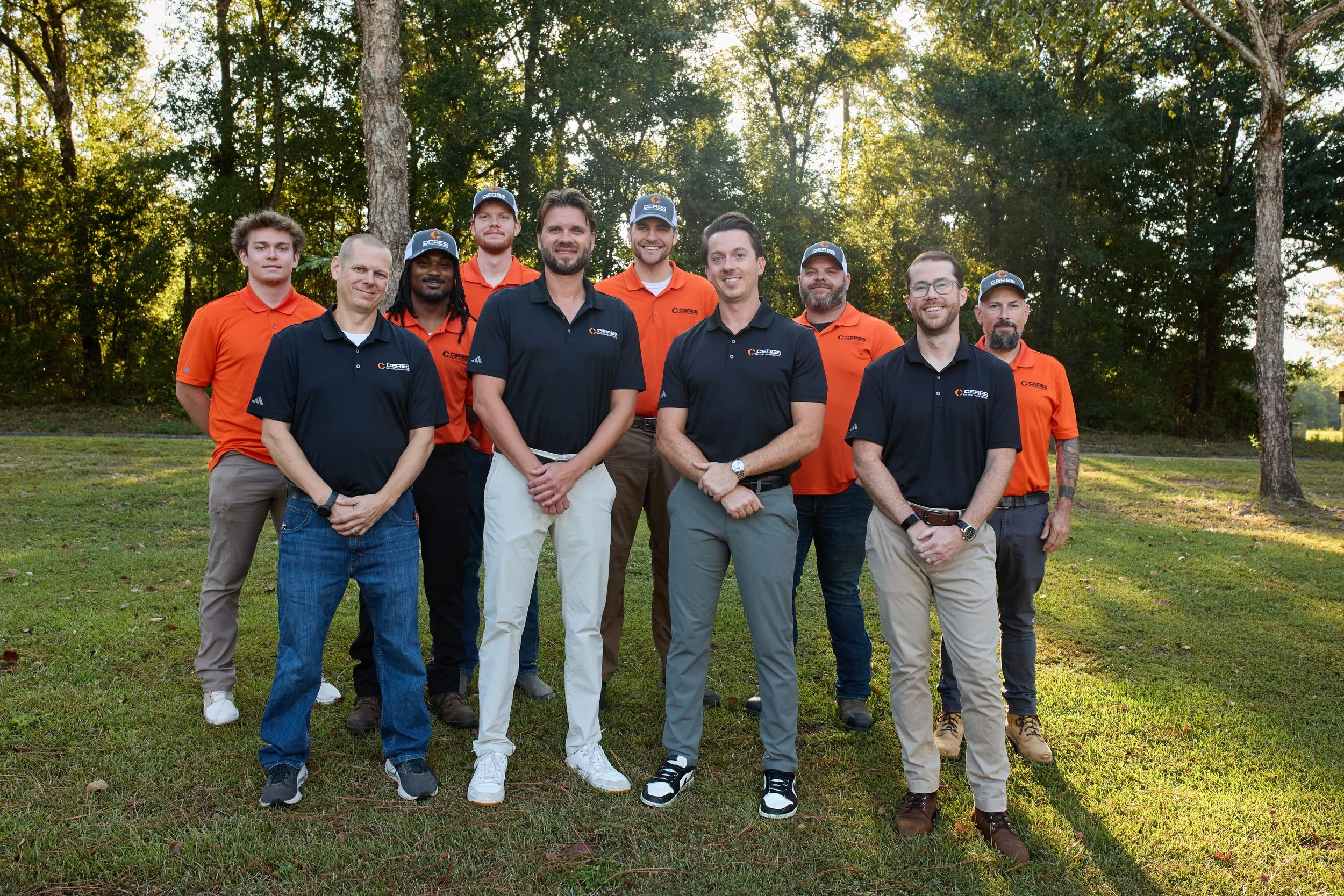 Group of eleven people standing outdoors on grass with trees in the background, some wearing black shirts and others in orange polo shirts, during the daytime